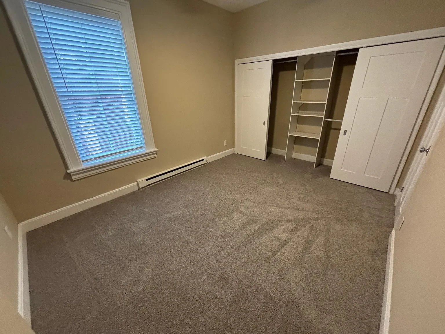 Empty bedroom with beige walls, one window with white blinds, beige carpet, white baseboards, and an open closet with double sliding doors and built-in shelving.