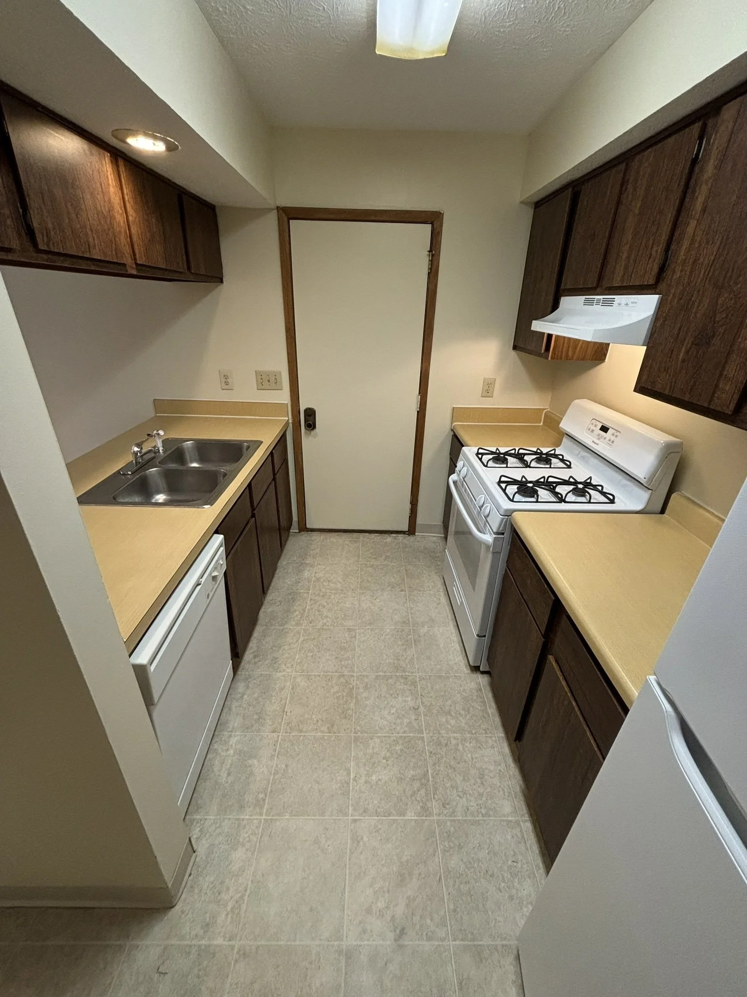 Small kitchen with dark wood cabinets, beige countertops, white appliances including a stove and refrigerator, double sink, and beige tiled floor.