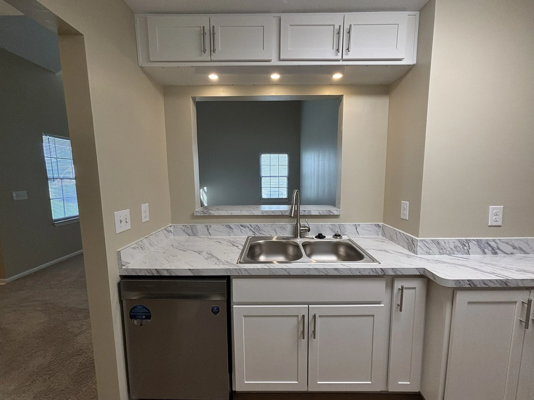 Kitchen with a granite countertop, double sink, white cabinets, small fridge, and a pass-through window to another room with a window.