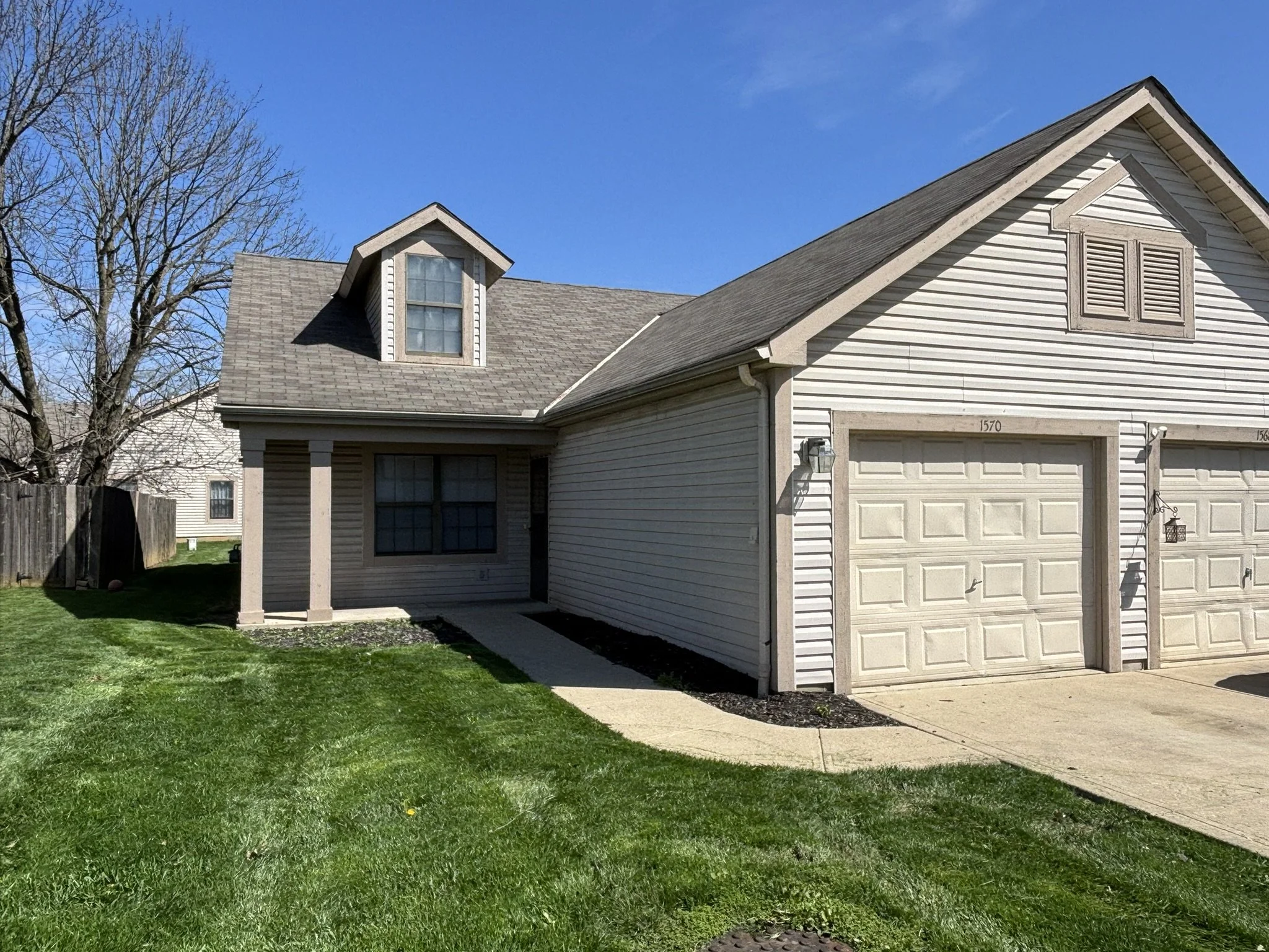 A two-story house with beige siding, a frontal garage, a porch with columns, and a second-story dormer window. The yard has green grass, and the sky is clear and blue.