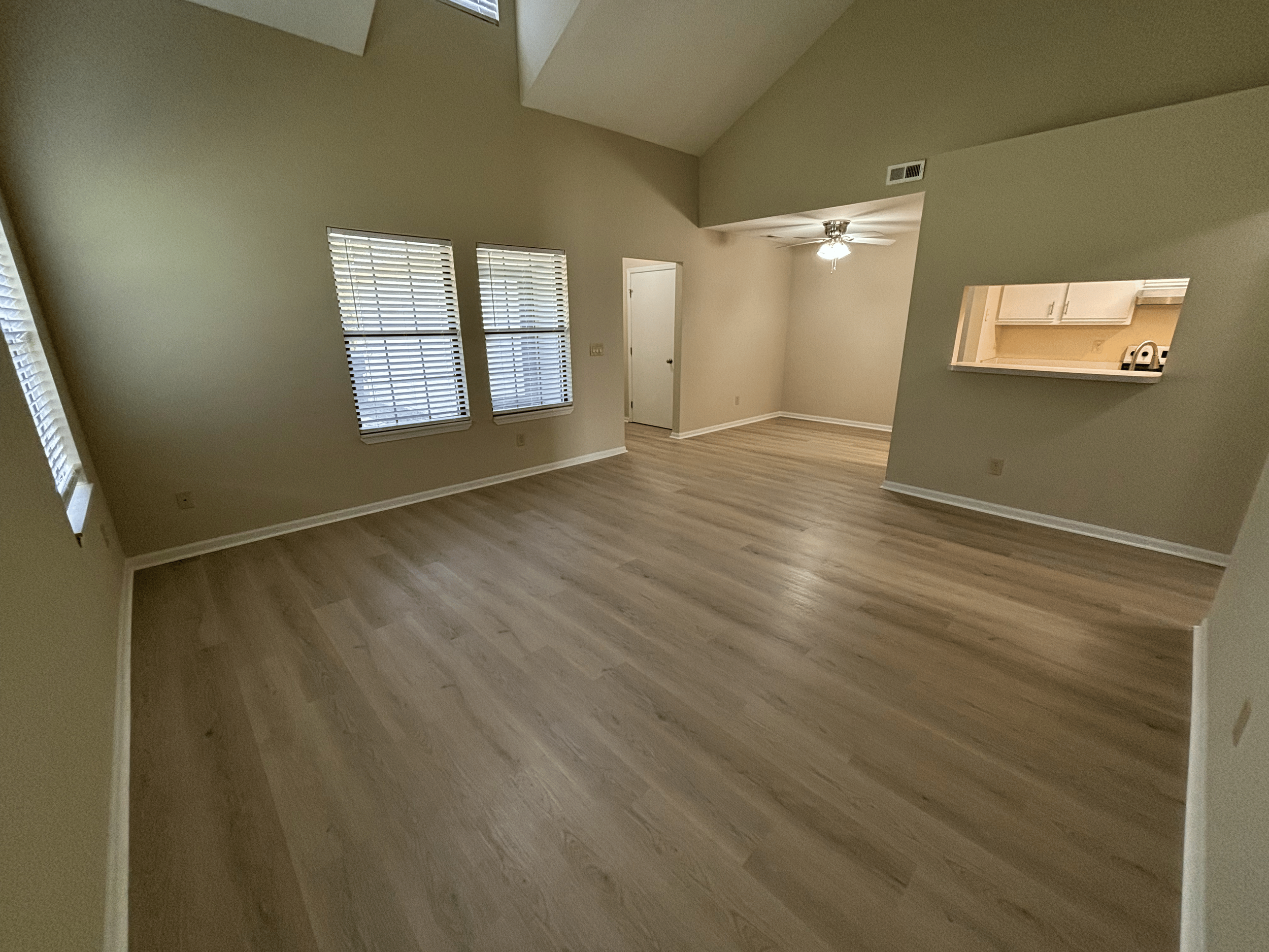 Empty living room with light-colored wooden floors, beige walls, three windows with blinds, a ceiling fan with light, and an open wall cutout to the kitchen.
