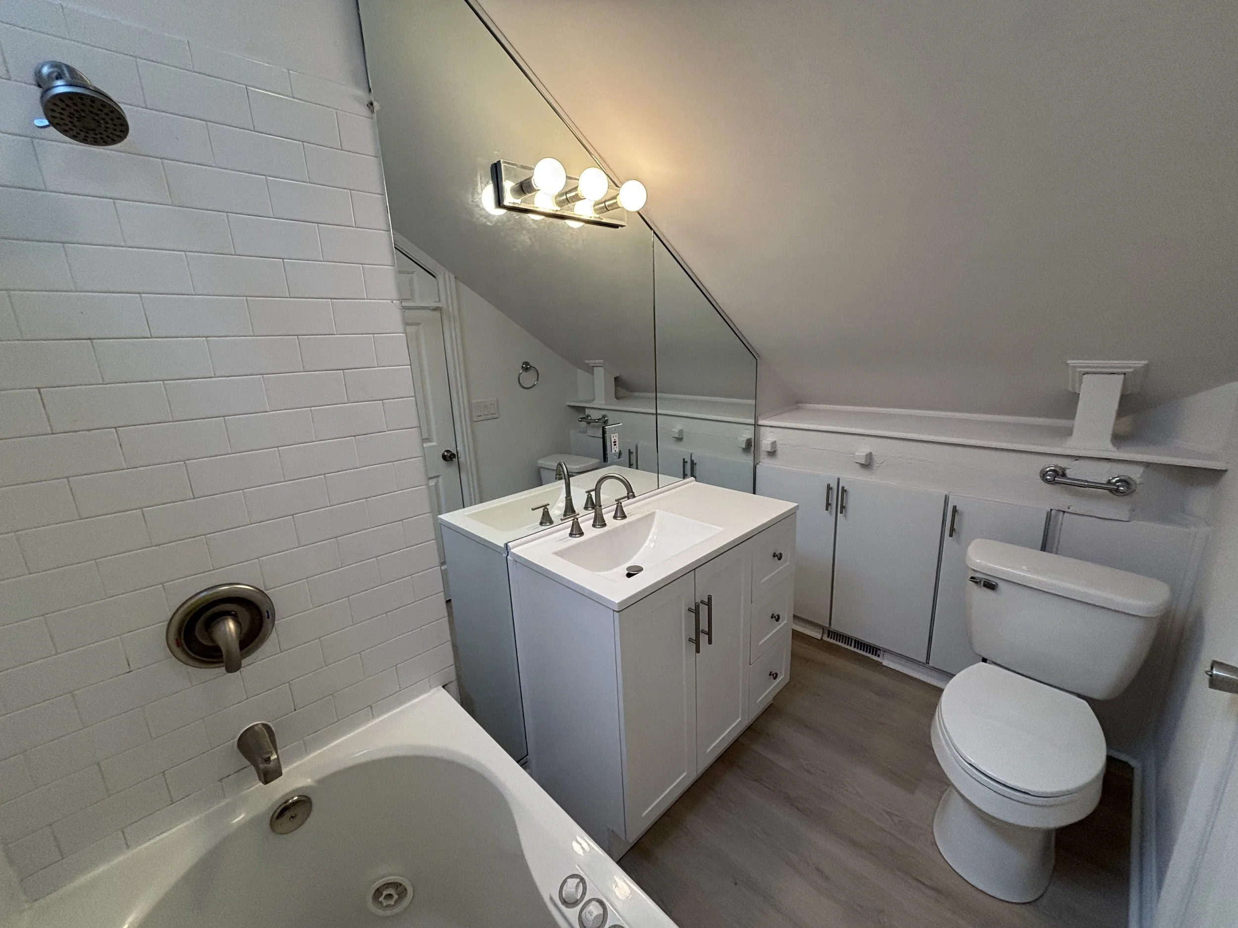 Bathroom with white tiled bathtub, white vanity with sink, mirror, storage cabinets, and toilet, under a sloped ceiling with a bright light fixture.