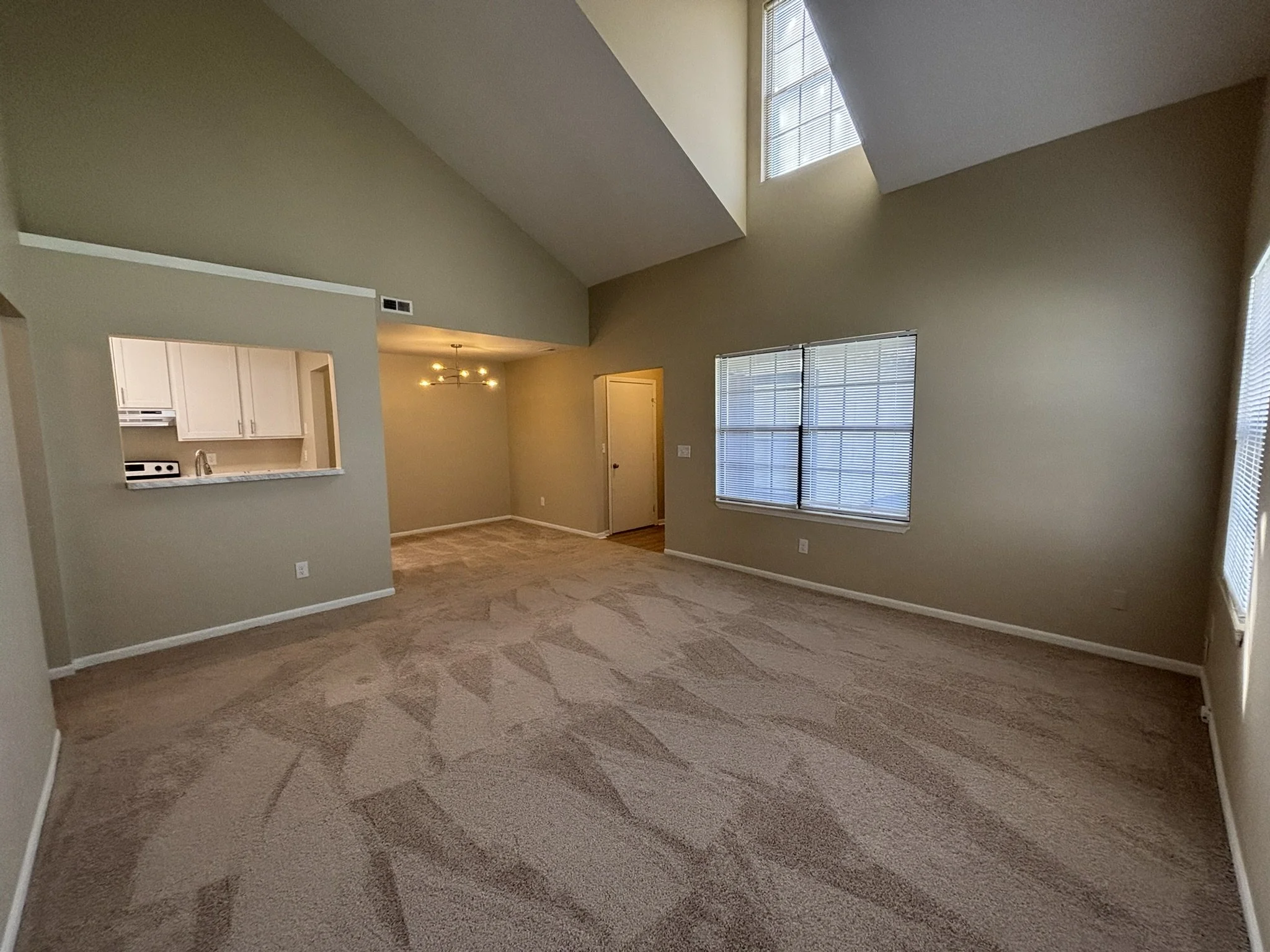 Empty living room with high vaulted ceiling, beige walls, large windows with blinds, and beige carpeted floors, with a view into the kitchen through a pass-through window.
