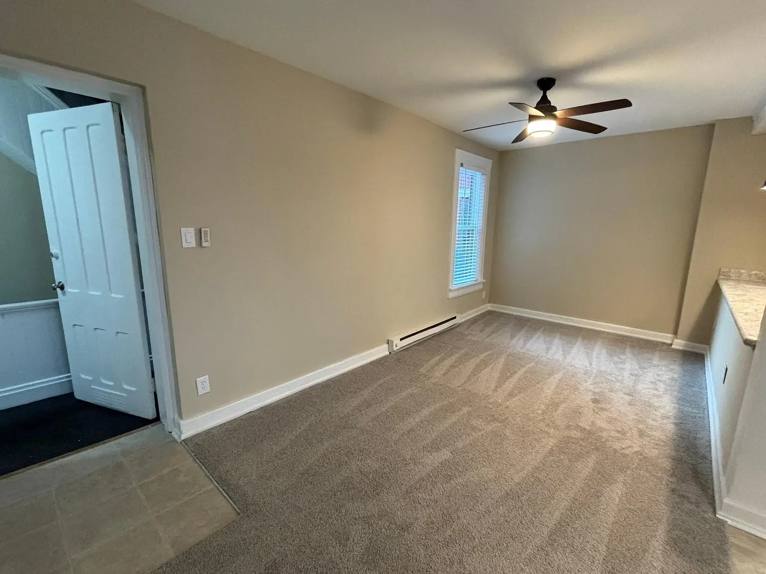 Empty living room with beige walls, carpeted floor, ceiling fan, and a window with blinds.