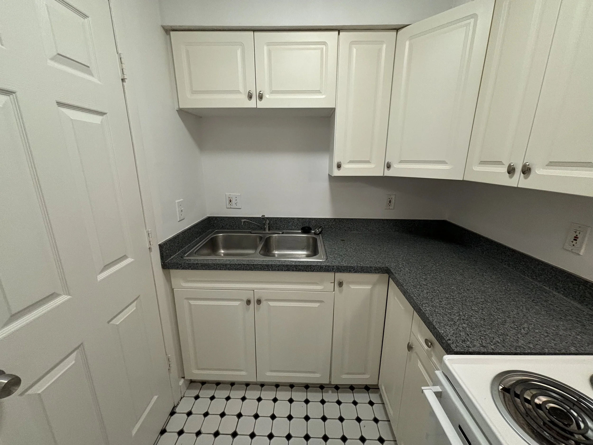 Kitchen with white cabinets, black countertop, double sink, electric stove, white walls, and black and white tiled floor.