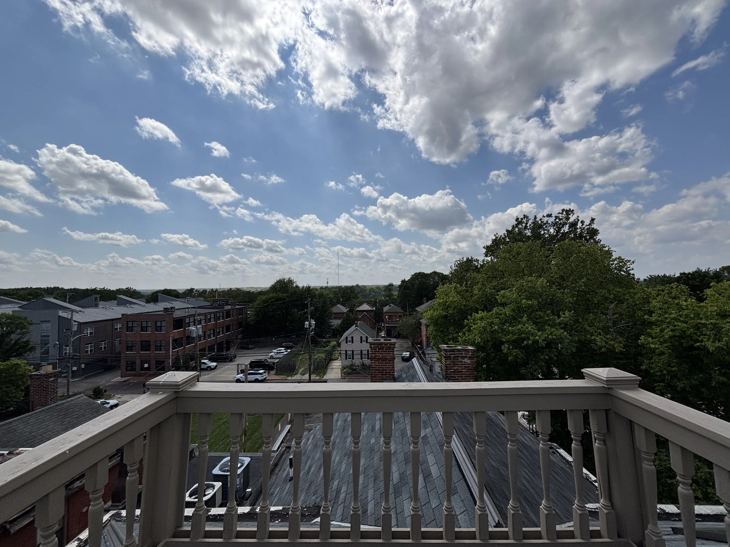 View from a balcony showing a mostly cloudy sky over a city neighborhood with apartment buildings, trees, cars, and rooftops.