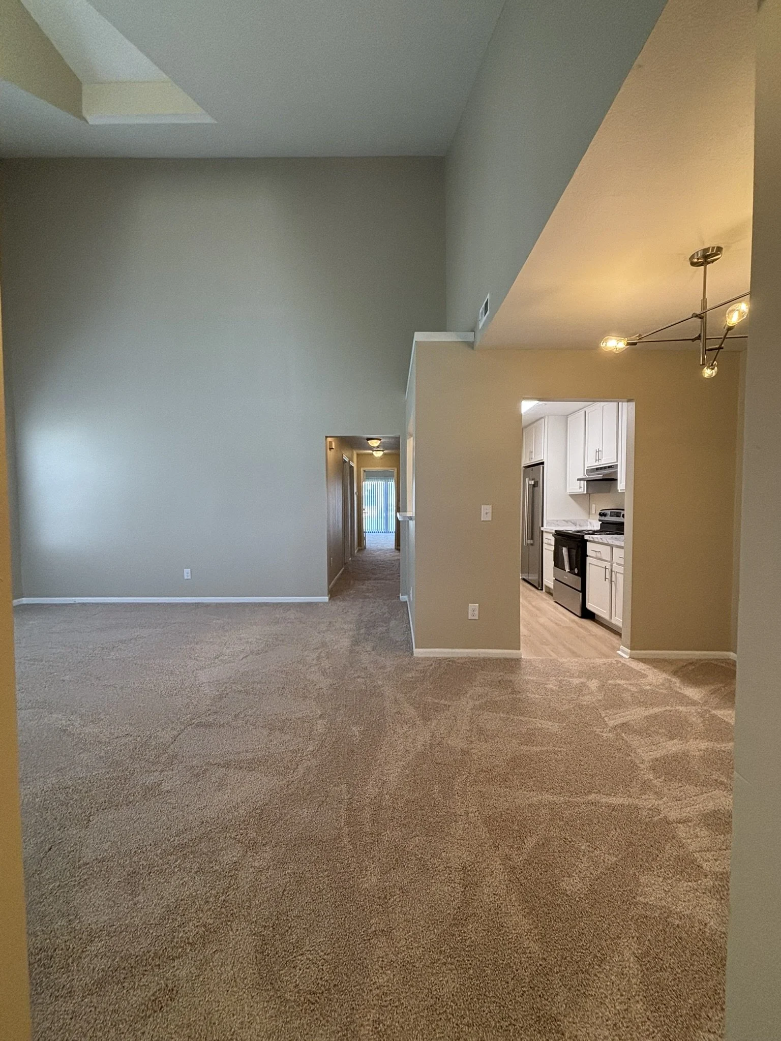 Empty living room with beige carpet and walls, view into adjacent kitchen with white cabinets and stainless steel appliances.