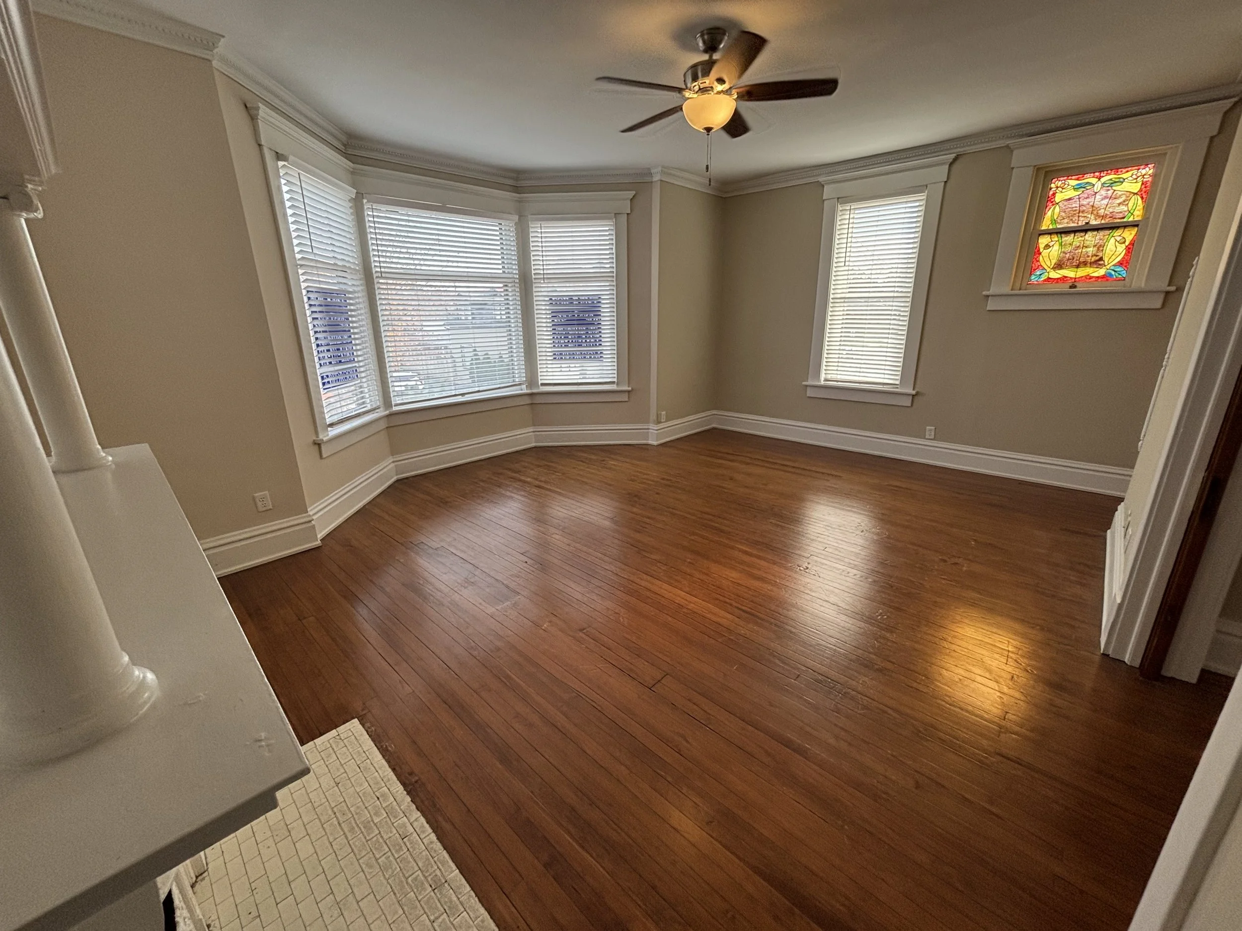 Empty living room with hardwood floors, three large windows with white blinds, a small stained glass window, a ceiling fan with light, and beige walls with white trim.
