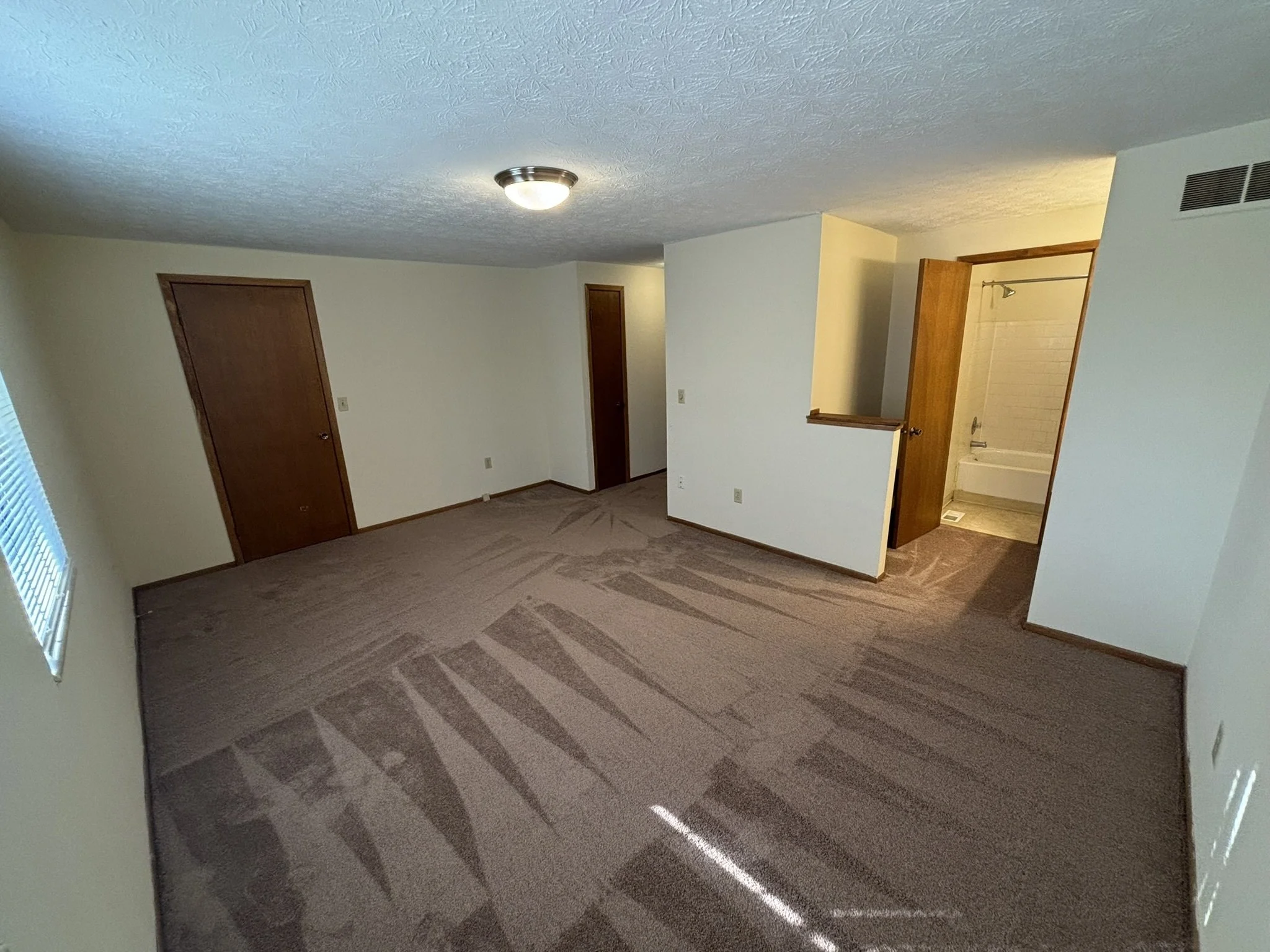 Empty living room with beige carpet, white walls, wooden doors, a window with blinds, and an adjacent bathroom with a bathtub.