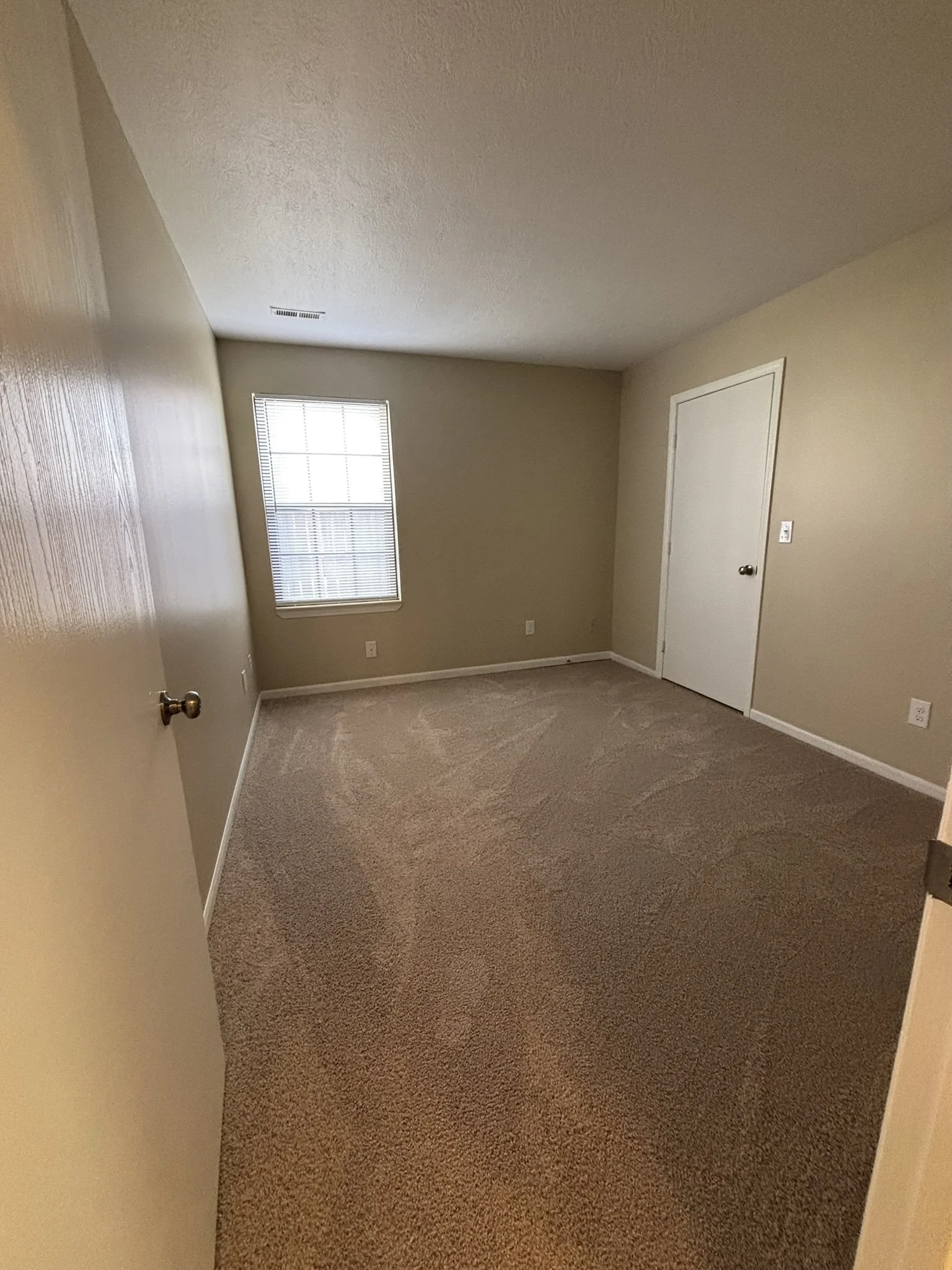 Empty bedroom with beige walls, a window with blinds, closed white door, and beige carpet.