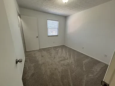 Empty bedroom with a window, a door, beige carpet, and plain white walls.