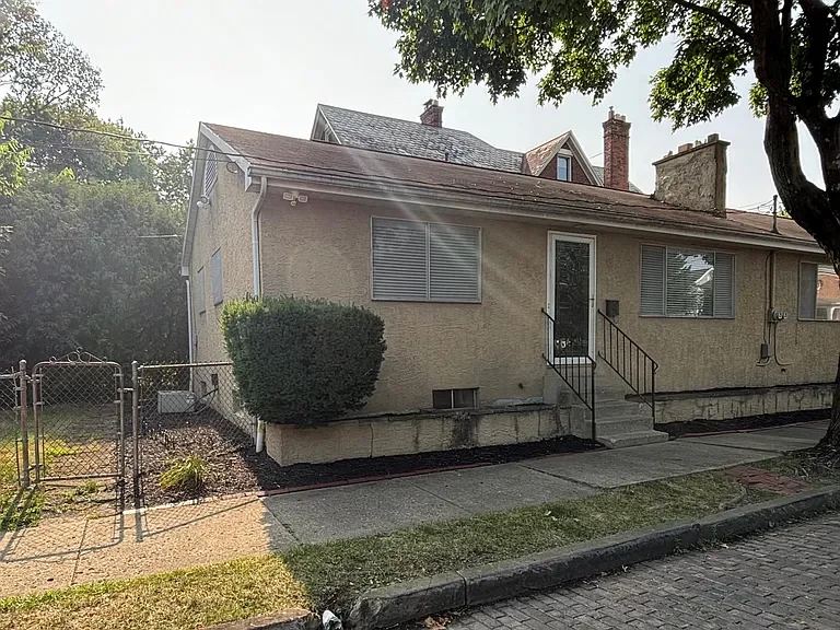 Front view of a beige house with a red roof, two front windows with blinds, a door accessible by a small staircase, a well-maintained front yard with a shrub, a sidewalk, and a tree providing shade.