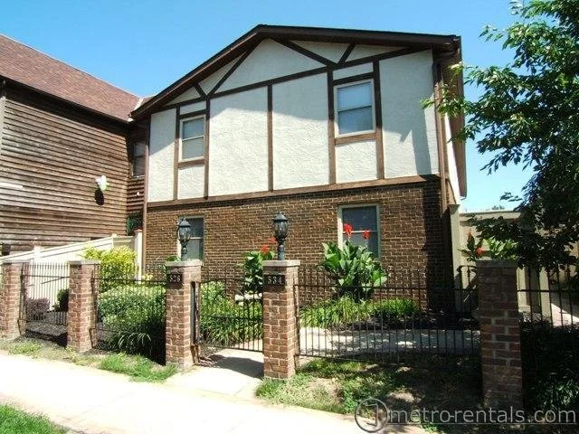 A two-story house with a tan stucco upper level and a brick lower level, surrounded by a brick fence with black metal gates and lamp posts, and a small garden with greenery and flowers.