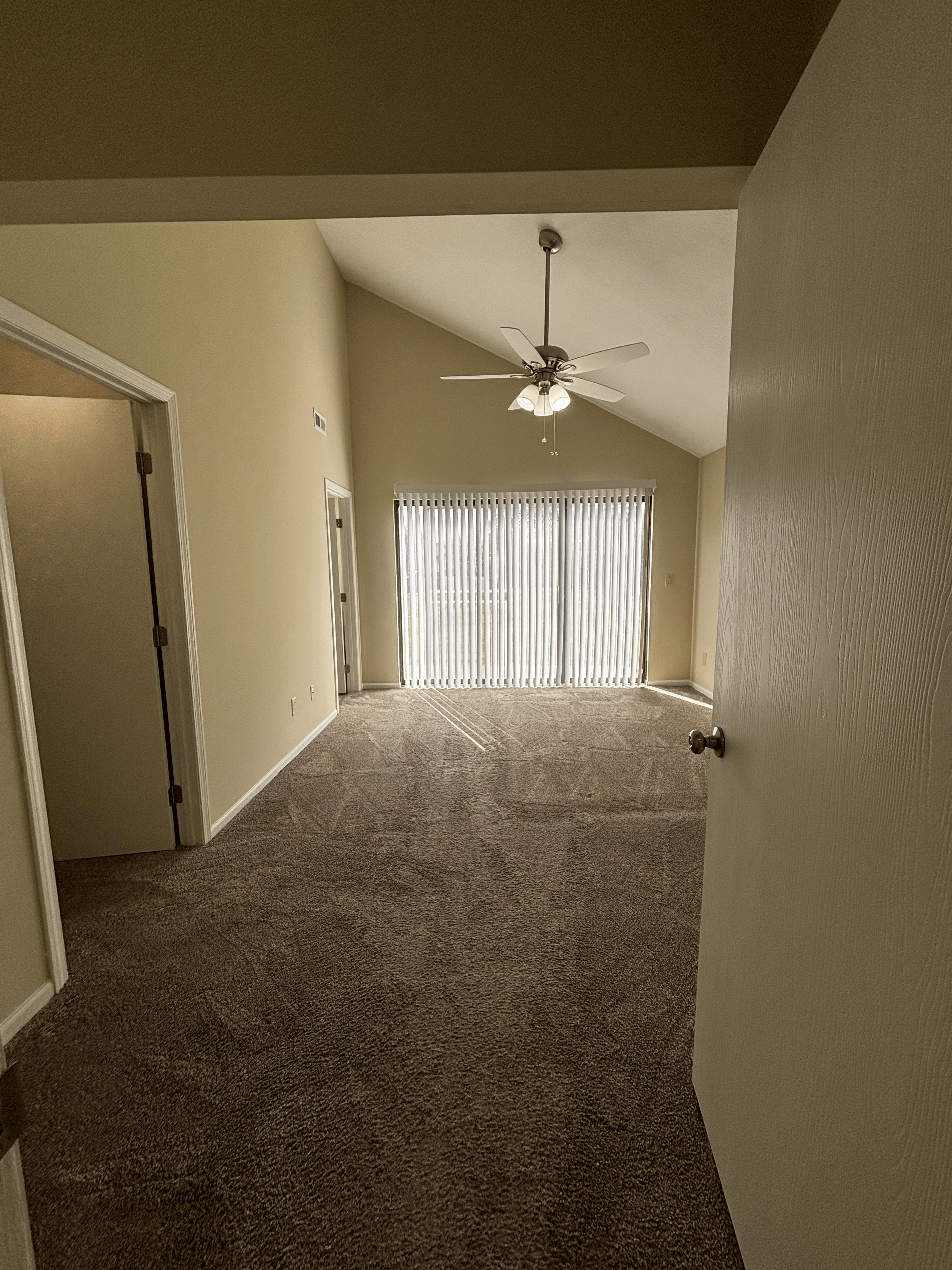 Empty living room with beige carpet, cream walls, a ceiling fan, vertical blinds covering a sliding glass door, and two closed doors on the left wall.