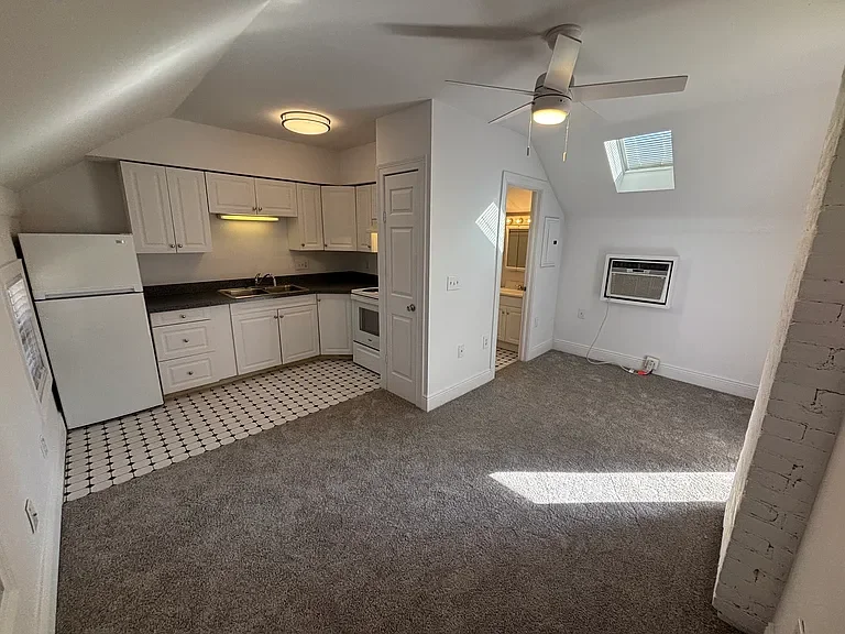 Empty living room and kitchen area in an apartment with white walls, gray carpet, a ceiling fan, a skylight, and an air conditioning window unit.