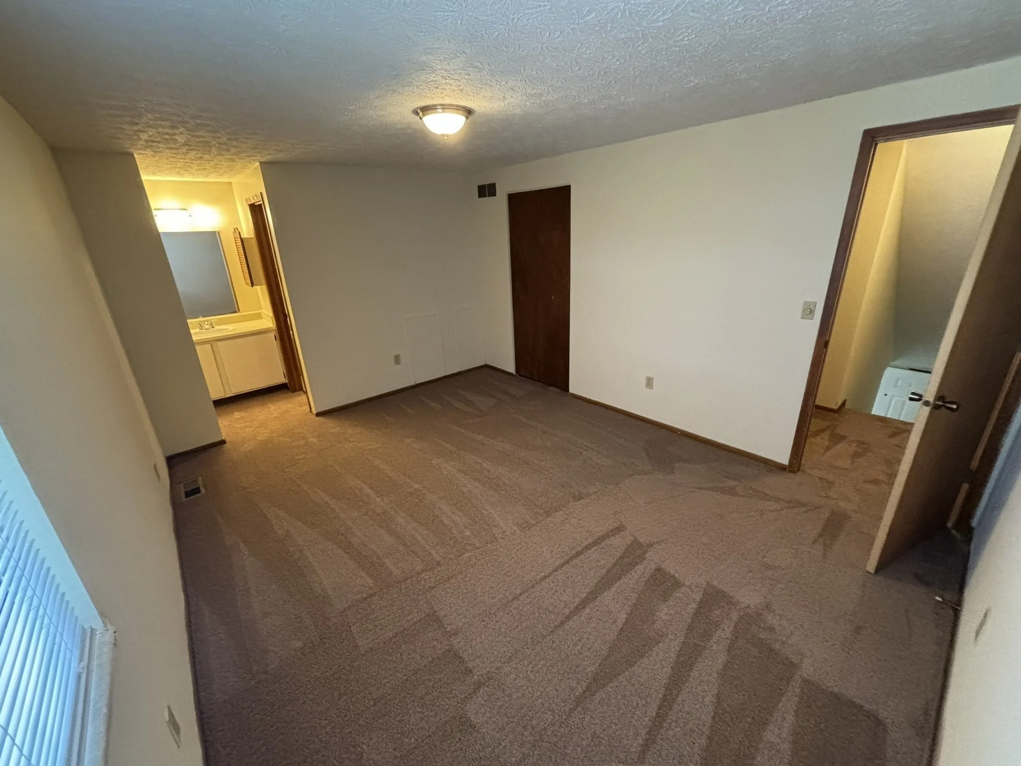 Empty living room with beige carpet and white walls, open door to small laundry room with white appliances, and a doorway leading to another room, with ceiling light fixture.