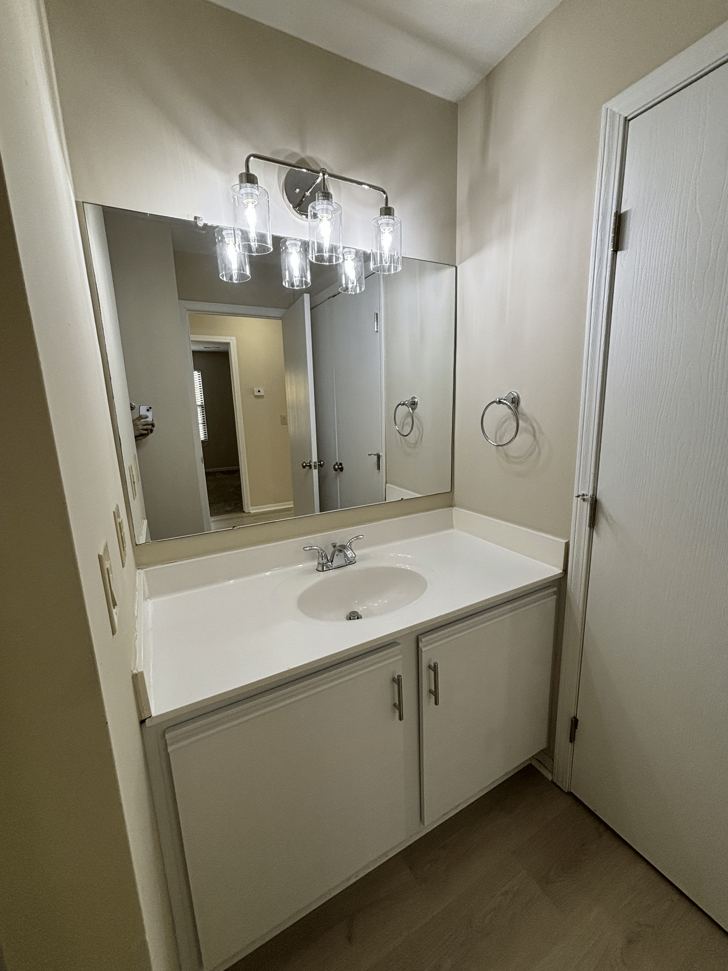 Bathroom vanity with a white countertop, sink, and cabinet doors beneath, a large mirror, chrome faucet, and light fixture with four glass shades, beige walls, and a door to the right.