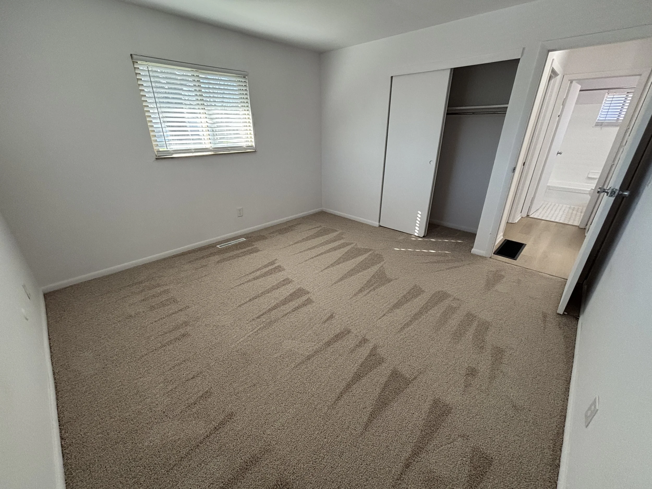 Empty bedroom with beige carpet, white walls, a window with blinds, an open closet with sliding doors, and a door leading to a bathroom.