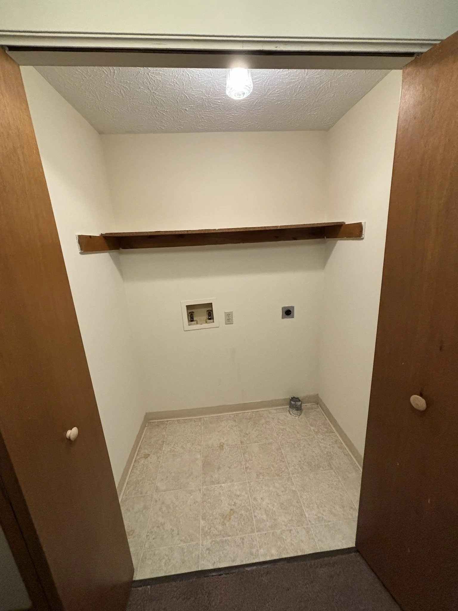 Empty laundry room with beige tiled floor, white walls, wooden cabinet doors, a wooden shelf, and washer/dryer hookups on the wall.