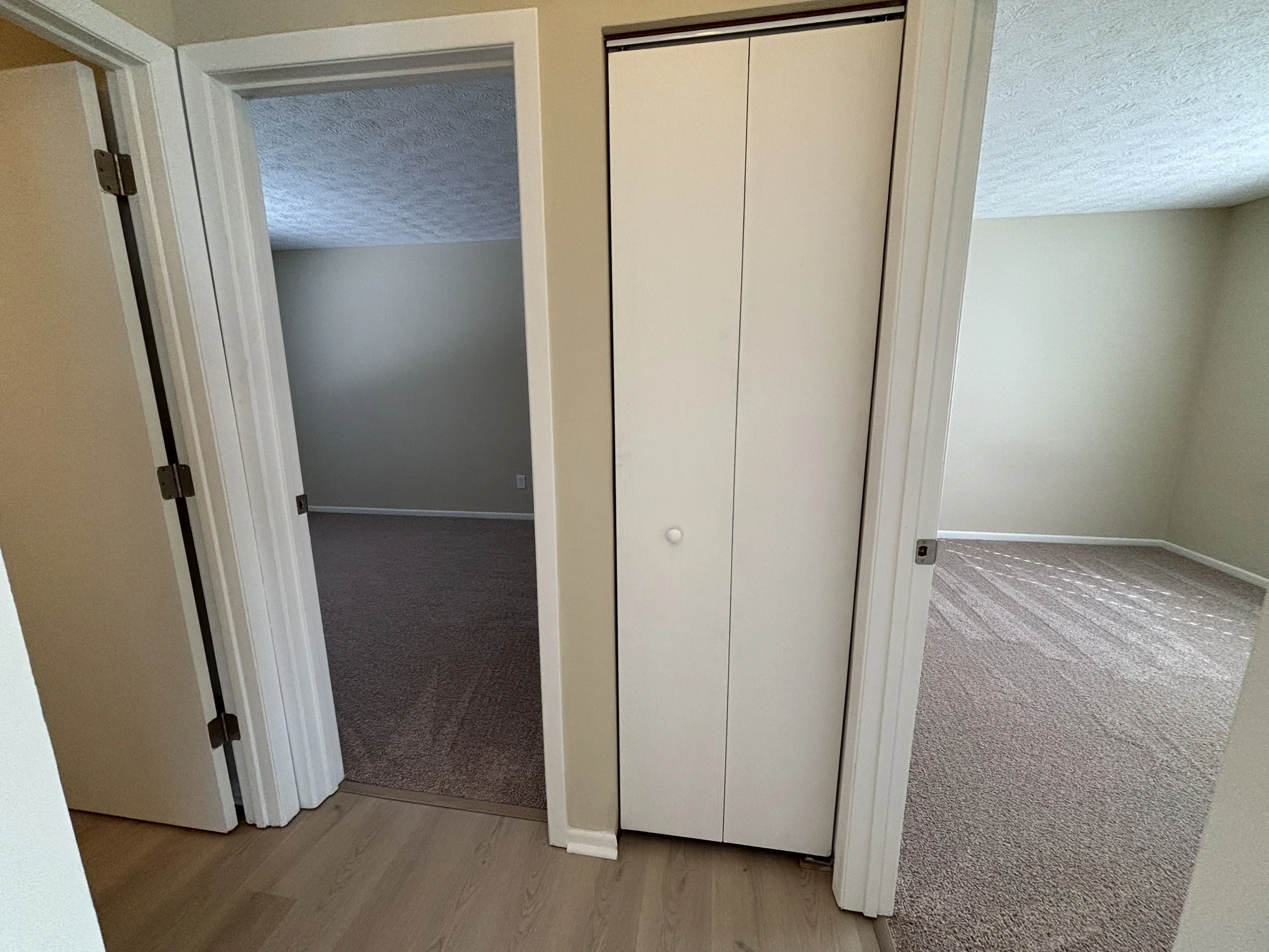 Interior view of a hallway with three rooms: one with closed white closet doors and two open doorways leading to carpeted rooms with beige walls and carpeting.