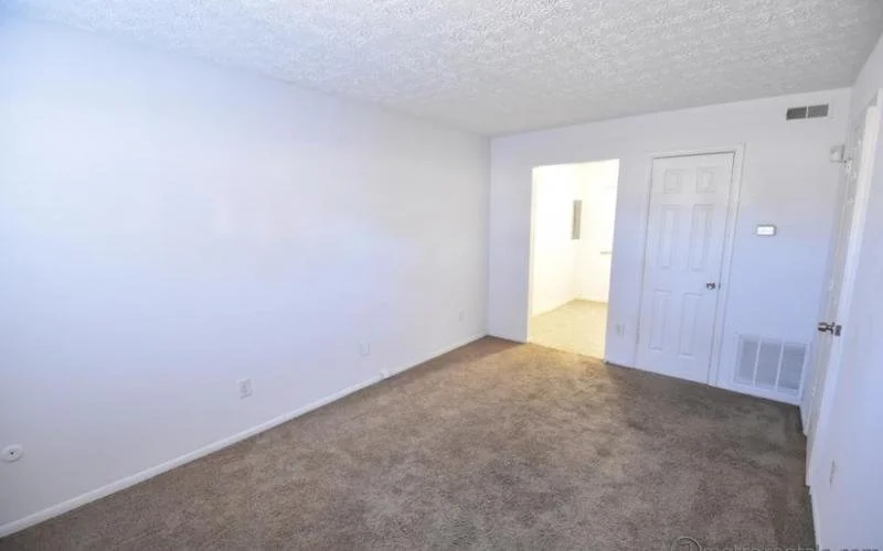 Empty living room with white walls, beige carpet, and doorways leading to other rooms.