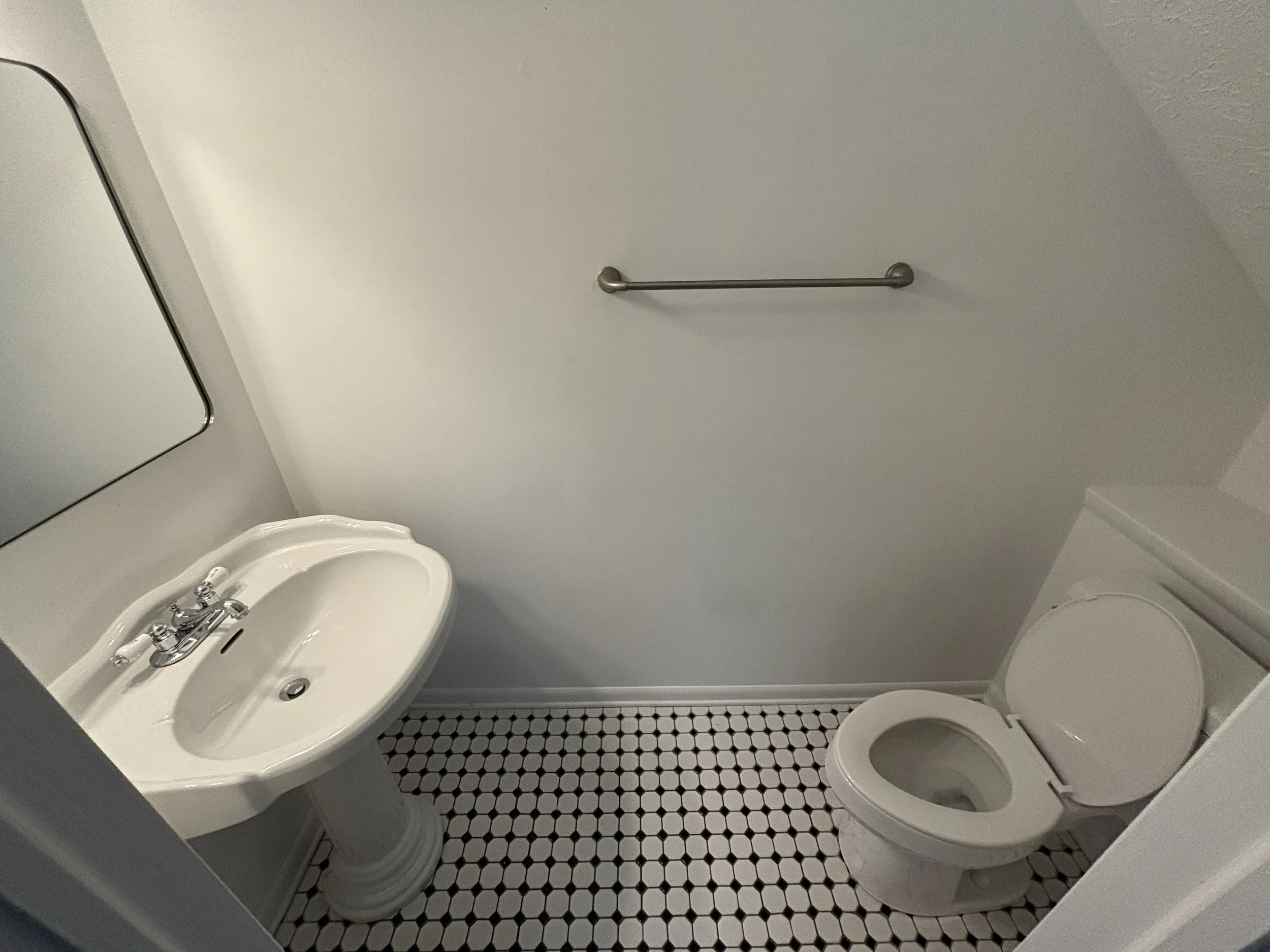 A small bathroom with a white pedestal sink, a mirror above it, and a toilet on the right. The wall is plain white with a towel bar mounted on it. The floor has small black and white hexagonal tiles.
