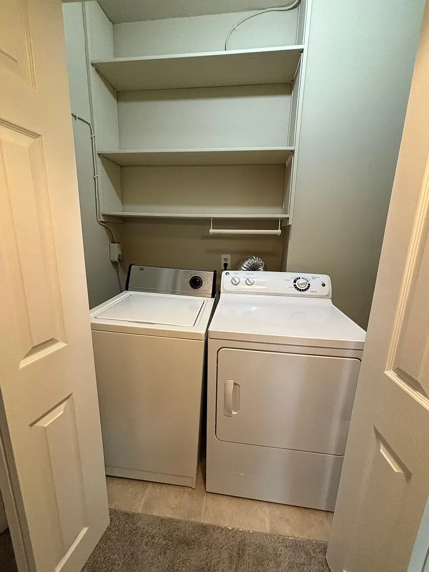 A small laundry room with a top-loading washing machine on the left and a front-loading dryer on the right. There are empty shelves above the appliances and a white rod underneath the middle shelf. The room has beige walls and a doorway with white do