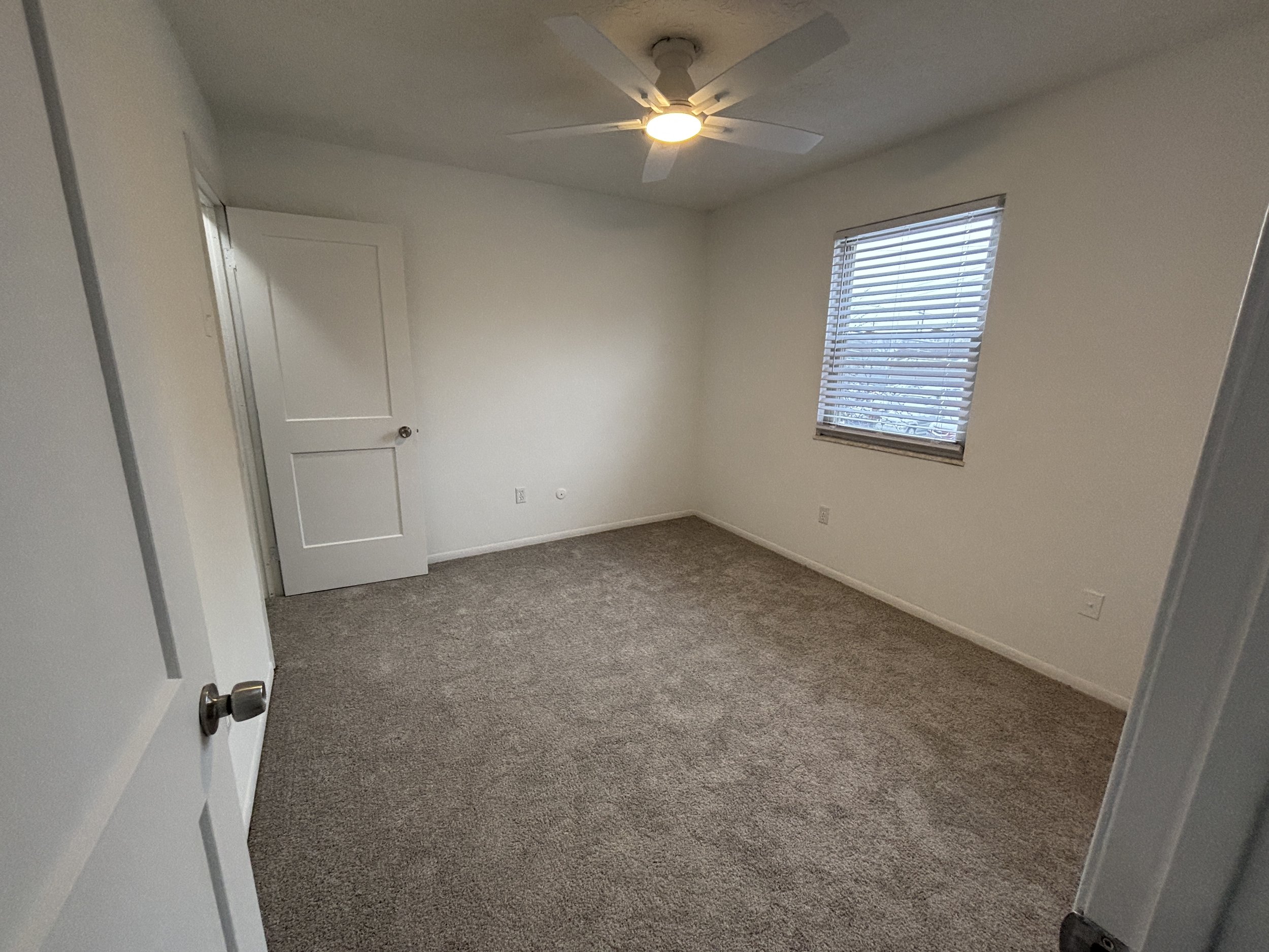 Empty bedroom with beige carpet, white walls, a window with blinds, a ceiling fan with a light, and a closed door.