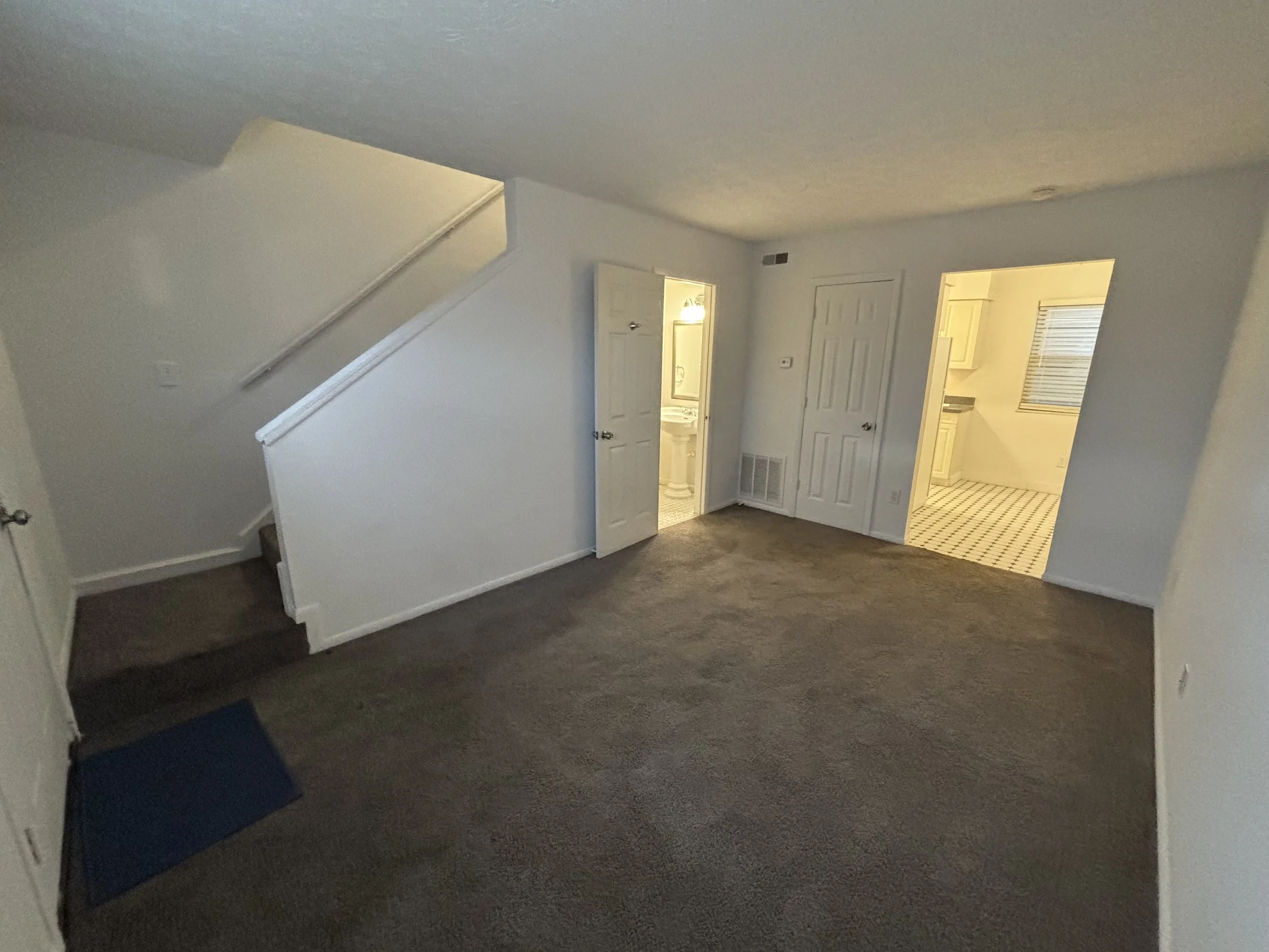 Empty living room with beige carpet, white walls, staircase on the left, three doors opening to a bathroom, closet, and kitchen, with a window in the kitchen area.