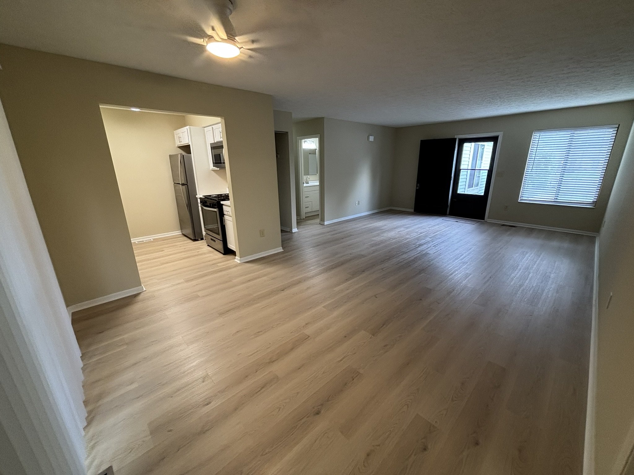 Empty living room with light wood flooring, beige walls, sliding glass door to balcony, window with blinds, and an open kitchen area.