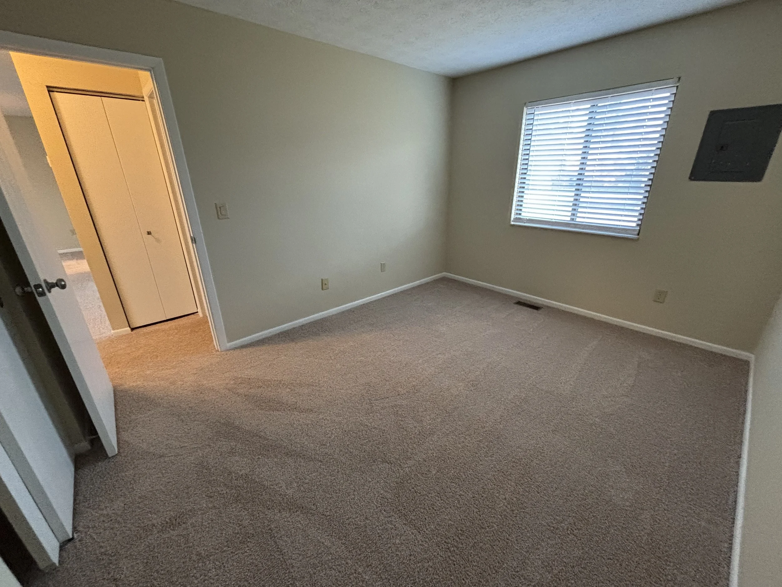 Empty bedroom with beige carpet, a window with white blinds, a closet with a sliding door, and a wall vent.
