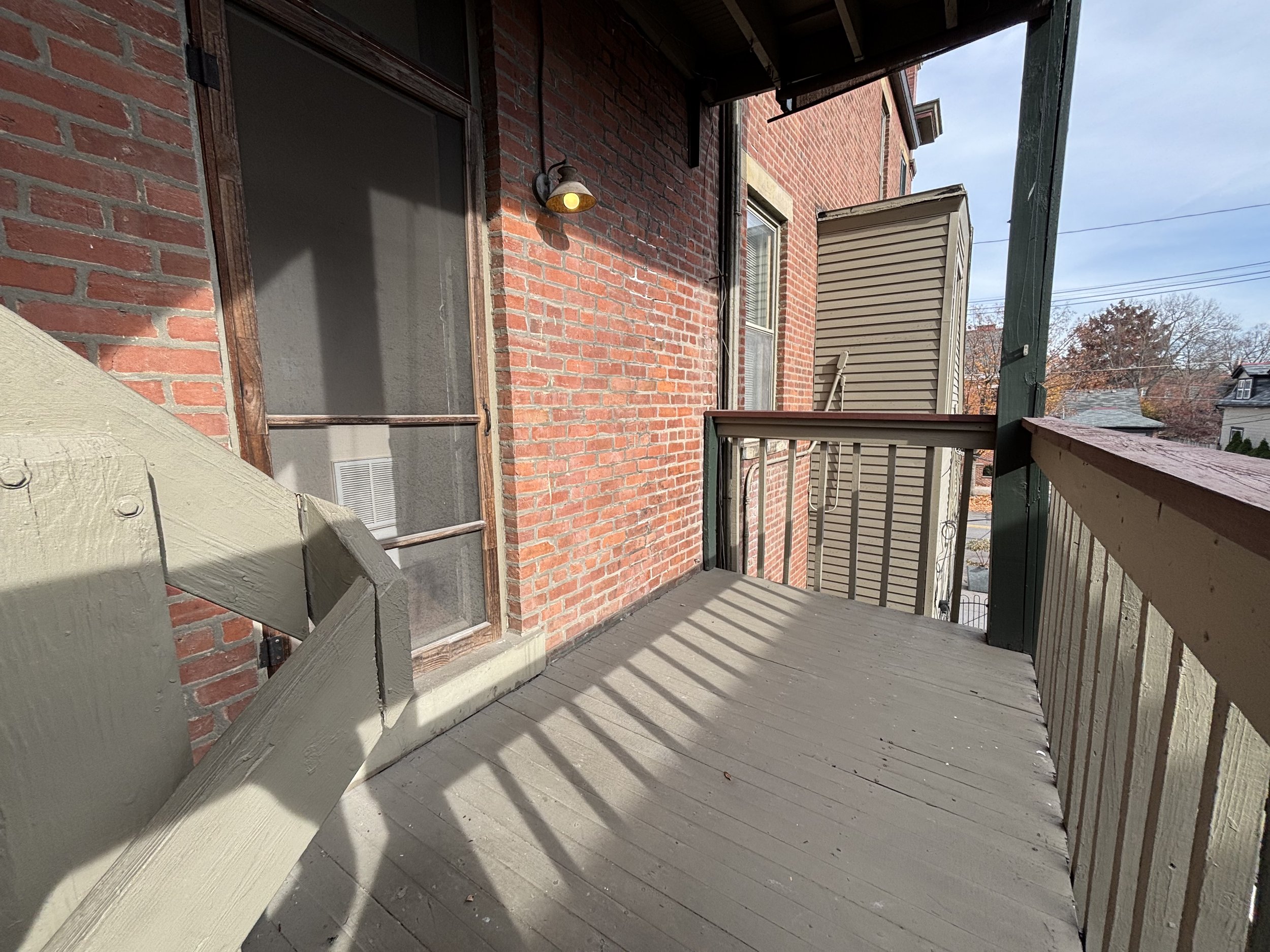 Small outdoor balcony with a wooden floor and railing, attached to a brick building, with a door and window, and overlooking a quiet residential neighborhood.