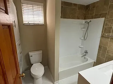 Bathroom with a toilet, window with blinds, and a shower/tub combo with a glass partition and built-in shelves.
