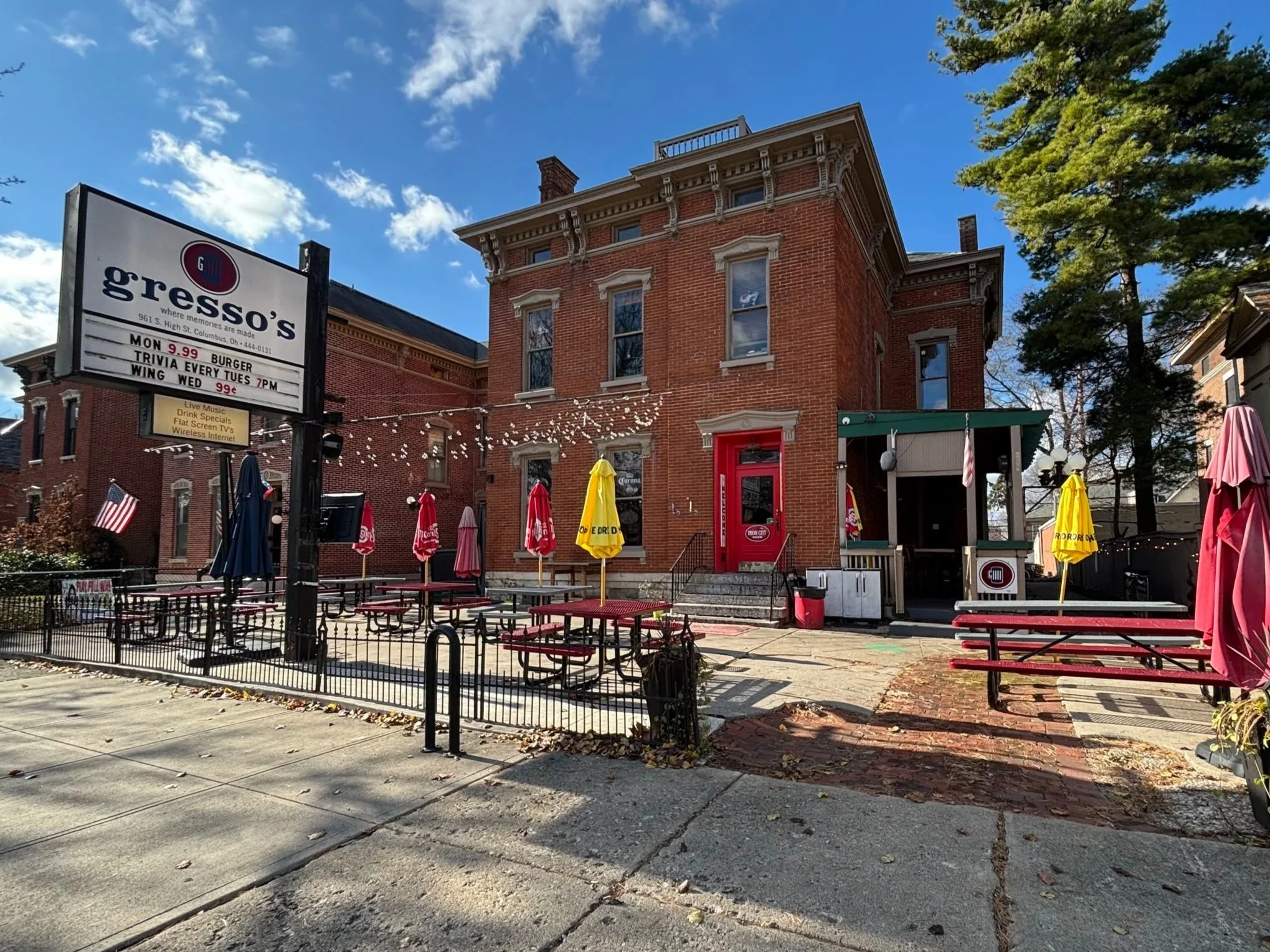 Exterior of a brick restaurant named Gresso's with outdoor patio tables, umbrellas, string lights, and a signboard, on a sunny day with a blue sky and trees in the background.