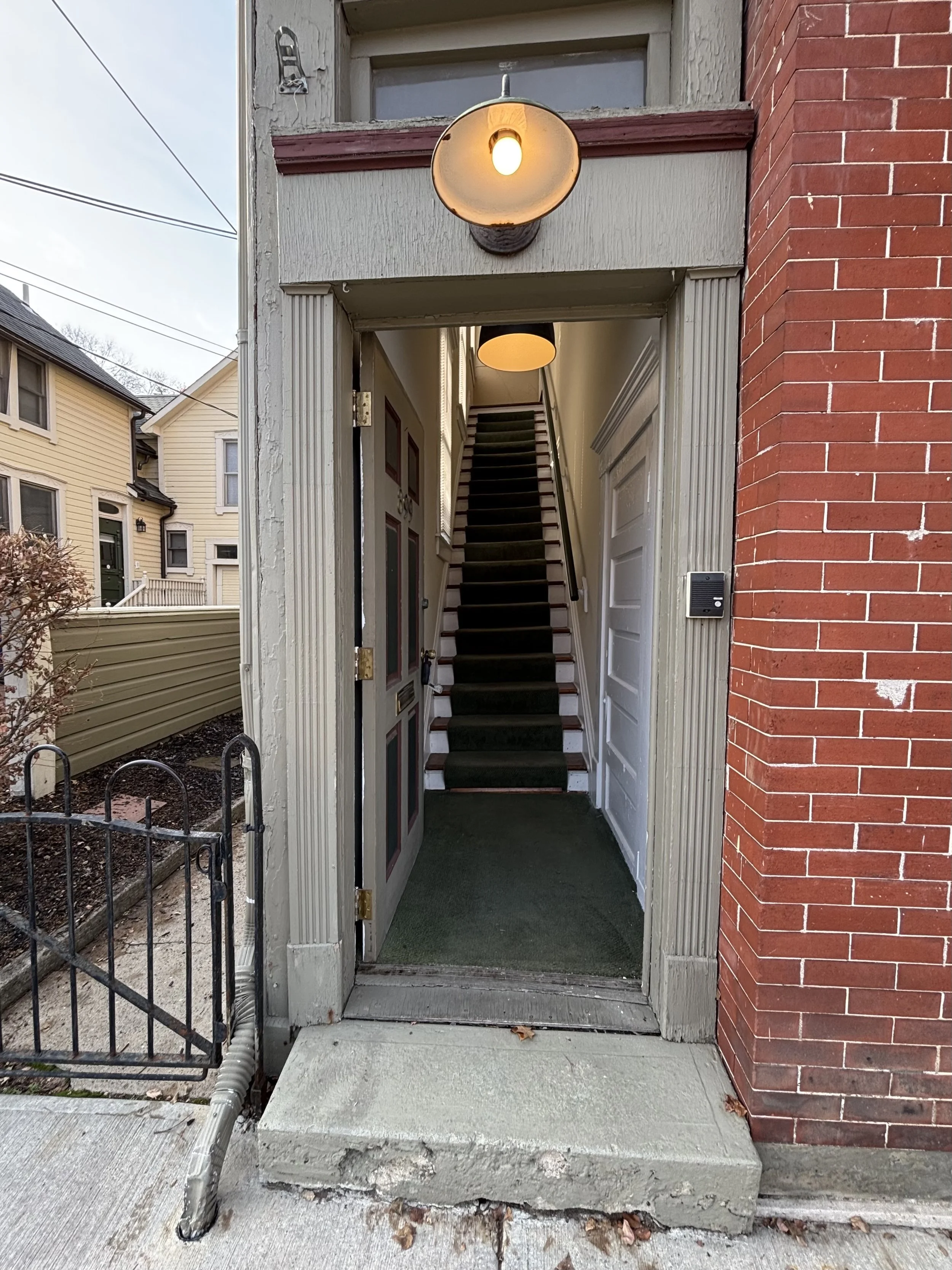 Entrance to a narrow staircase in an urban residential building with a door, carpeted stairs, and a wall-mounted light fixture.