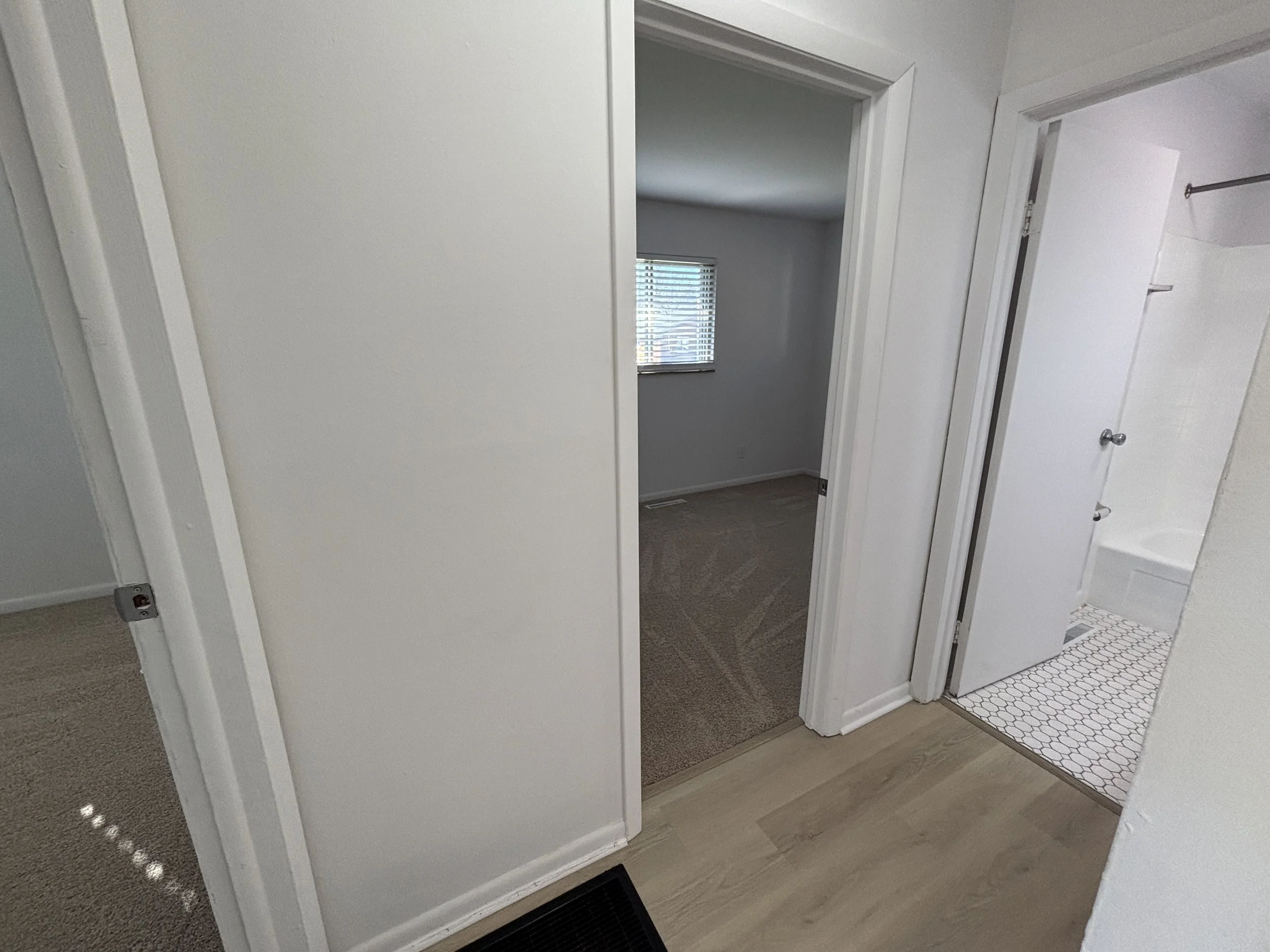 View of an entryway leading to a bedroom and bathroom with white walls, light wood floor, beige carpet in bedroom, and white tiled bathroom floor.