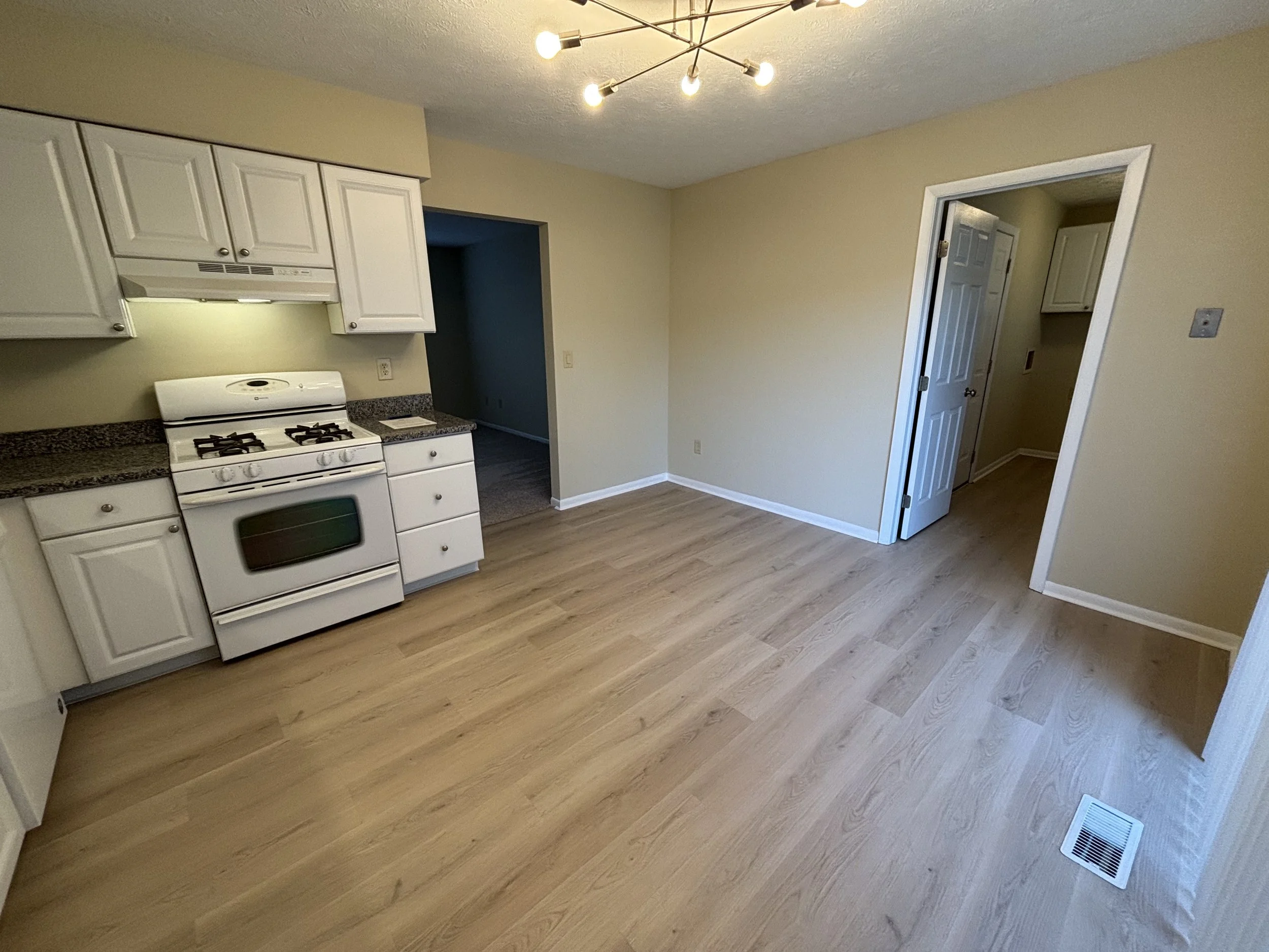 Empty kitchen with white cabinets, a white stove, and light wood flooring. An open doorway leads to another room, and there is a modern ceiling light fixture.
