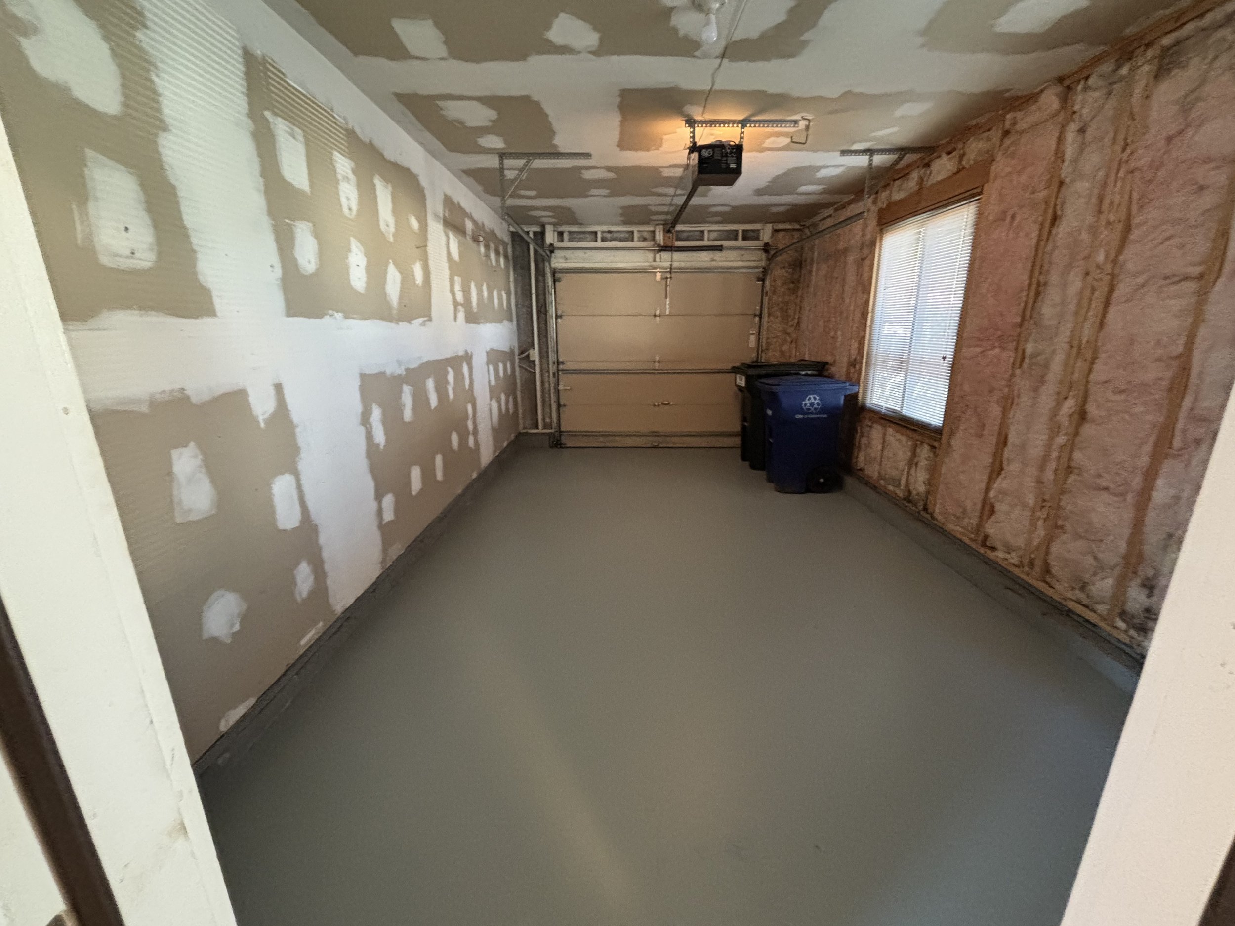 Empty garage with unfinished walls, a garage door, and a window with blinds. There are trash bins in the corner.