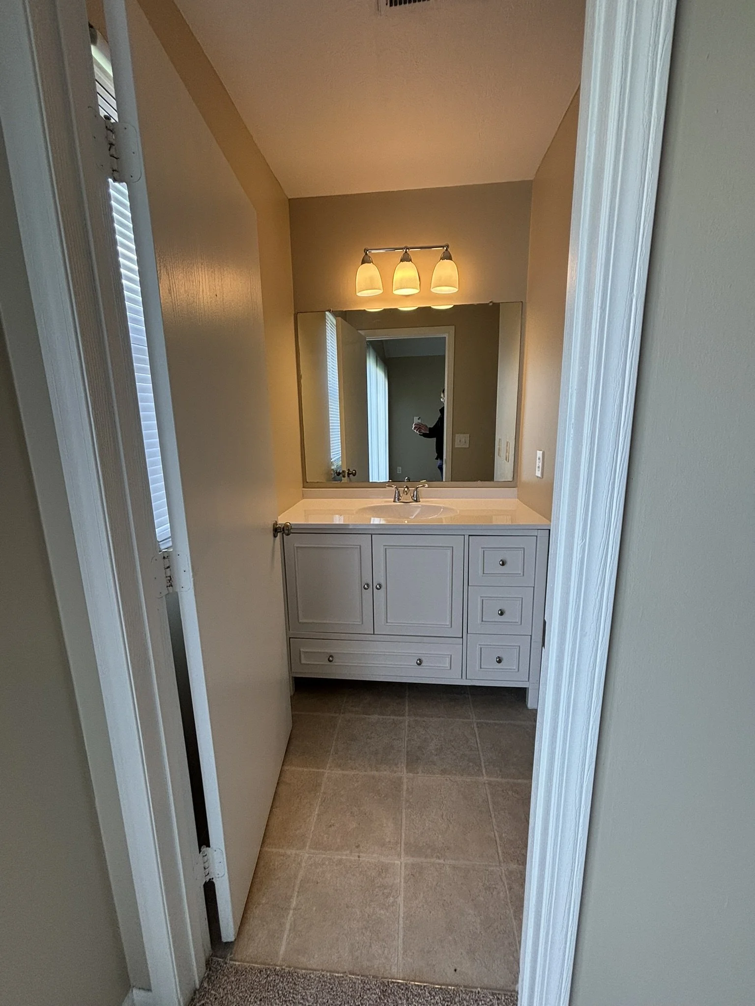 A bathroom with a white vanity, a large mirror, three light fixtures, and beige wall paint. Part of a window with blinds is visible on the left side.