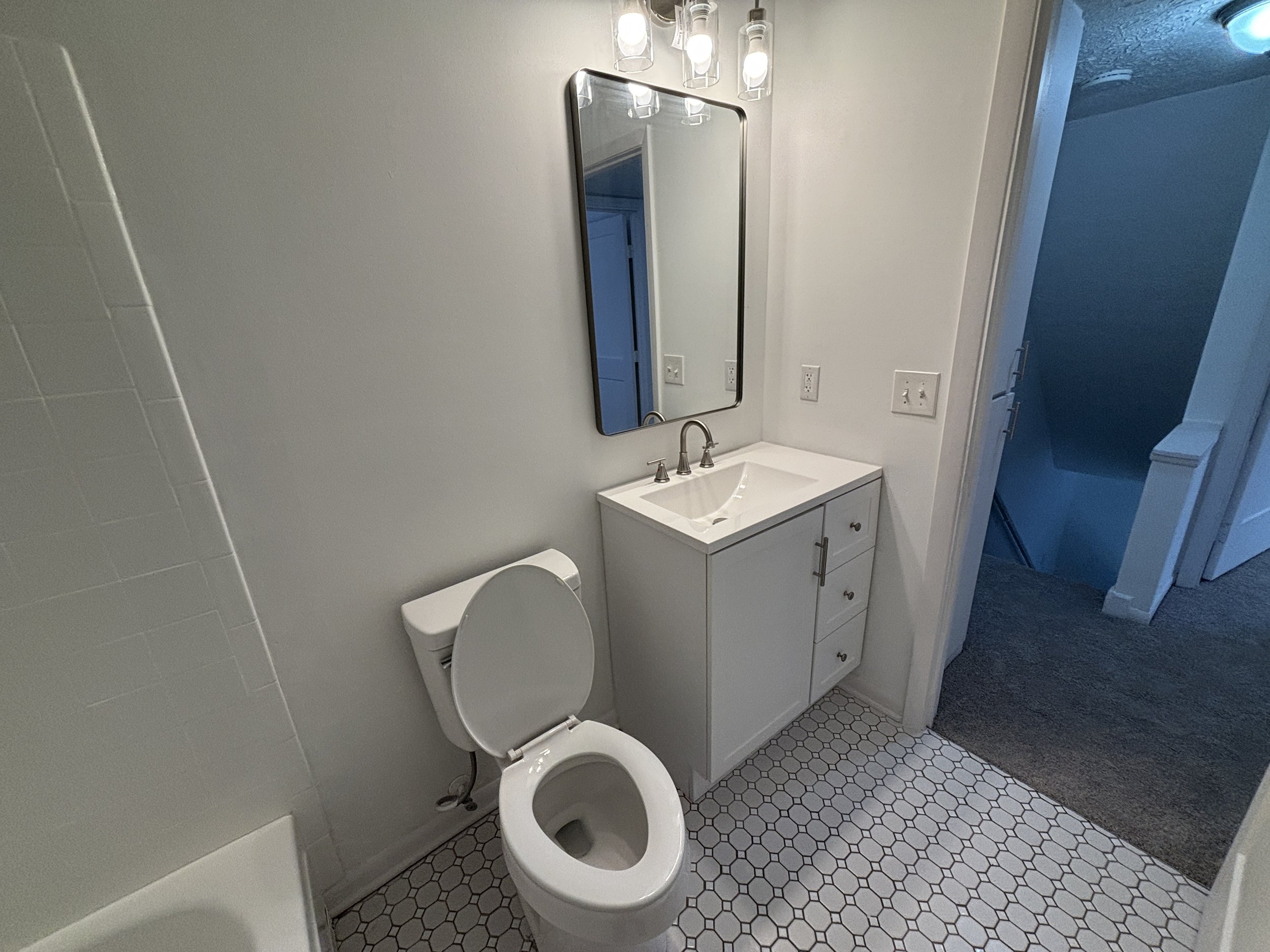 Bathroom with a white toilet, small white vanity with a sink, mirror above the sink, and wall-mounted light fixtures. The floor has hexagonal tiles, and there is a doorway leading to a carpeted room.