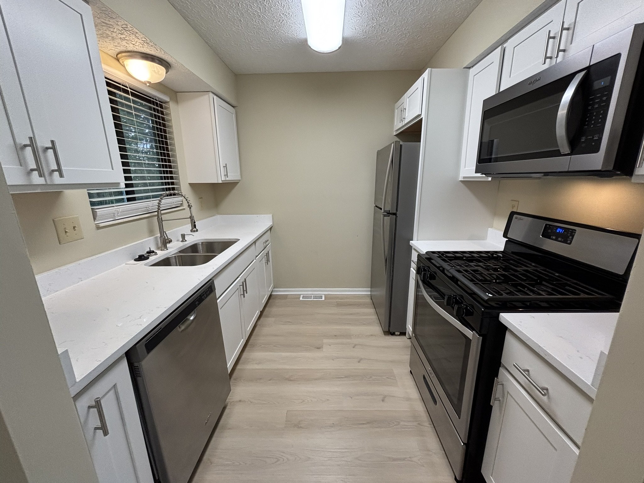 Kitchen with white cabinets, stainless steel appliances including a microwave, oven, refrigerator, and dishwasher, with a window above the sink and light-colored wood flooring.