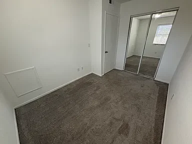 Empty bedroom with beige carpet, white walls, a mirrored closet with sliding doors, and a window providing natural light.