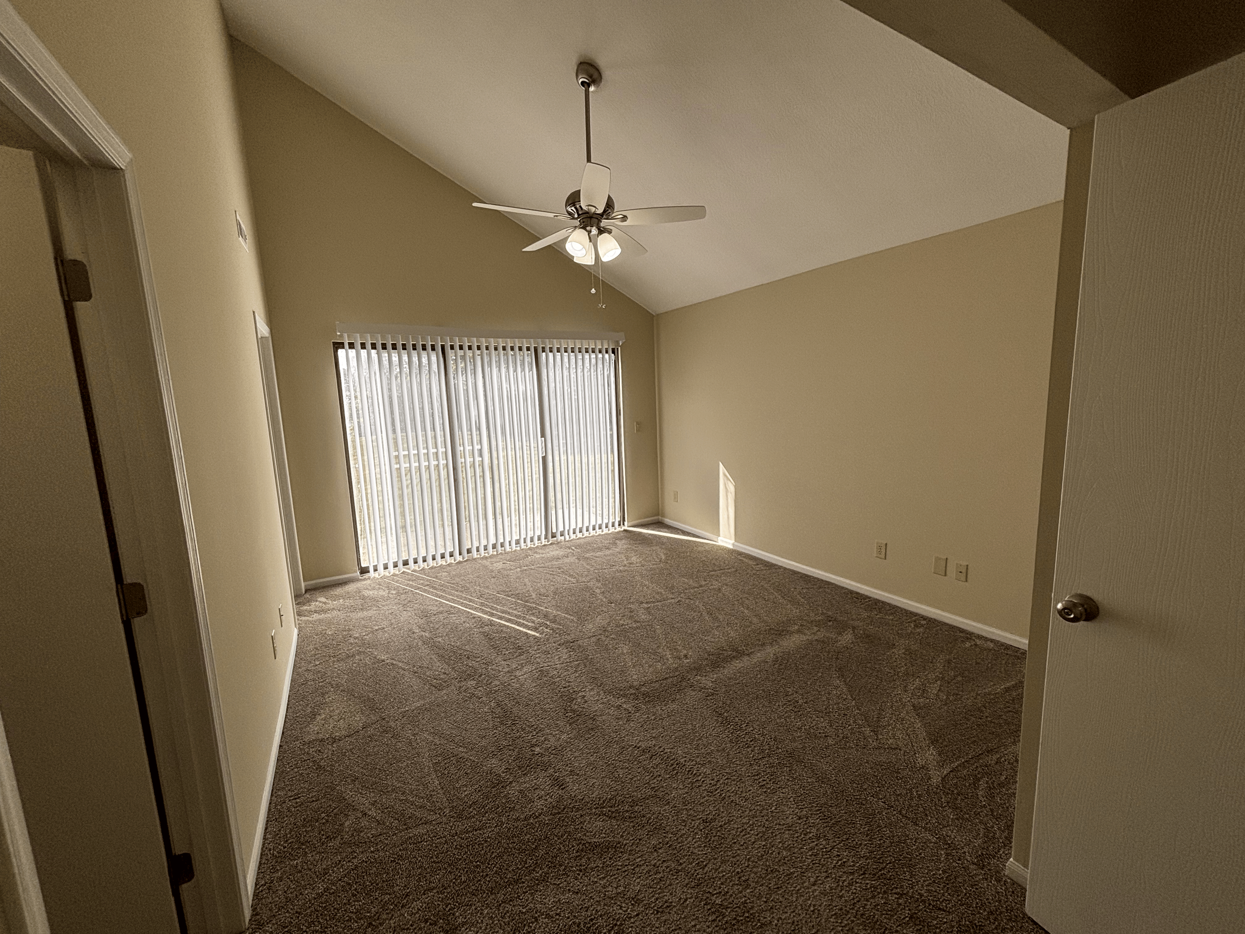 Empty living room with beige walls, a ceiling fan, carpeted floor, and sliding glass doors with vertical blinds.