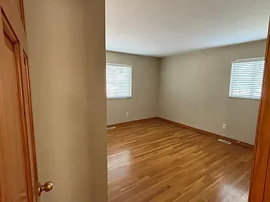 Empty room with hardwood floors, beige walls, and two windows with blinds.