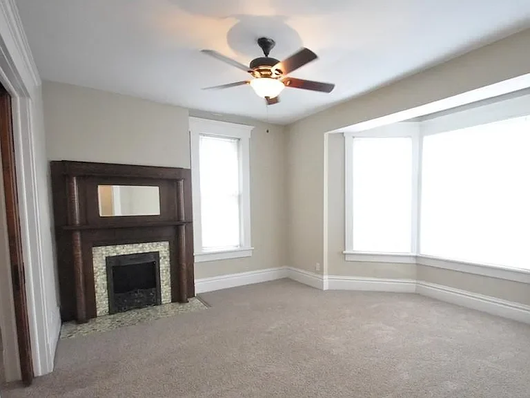 Empty living room with beige walls, carpeted floor, a fireplace with a wooden mantle, two windows, and a ceiling fan.