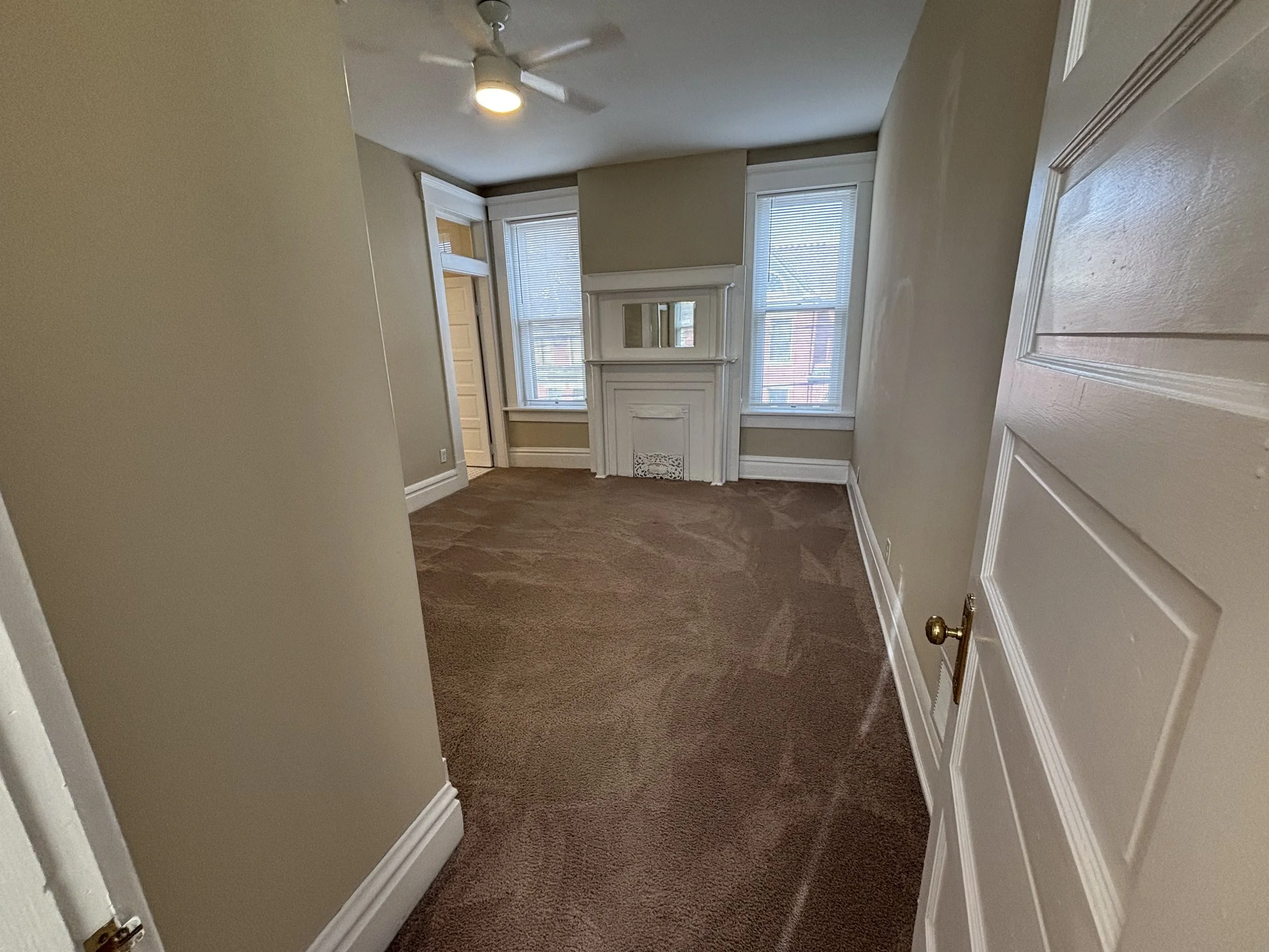 Empty room with beige walls, brown carpet, white baseboards, a white door on the right, three windows with blinds, a small fireplace with a mirror above, and a ceiling fan with a light.