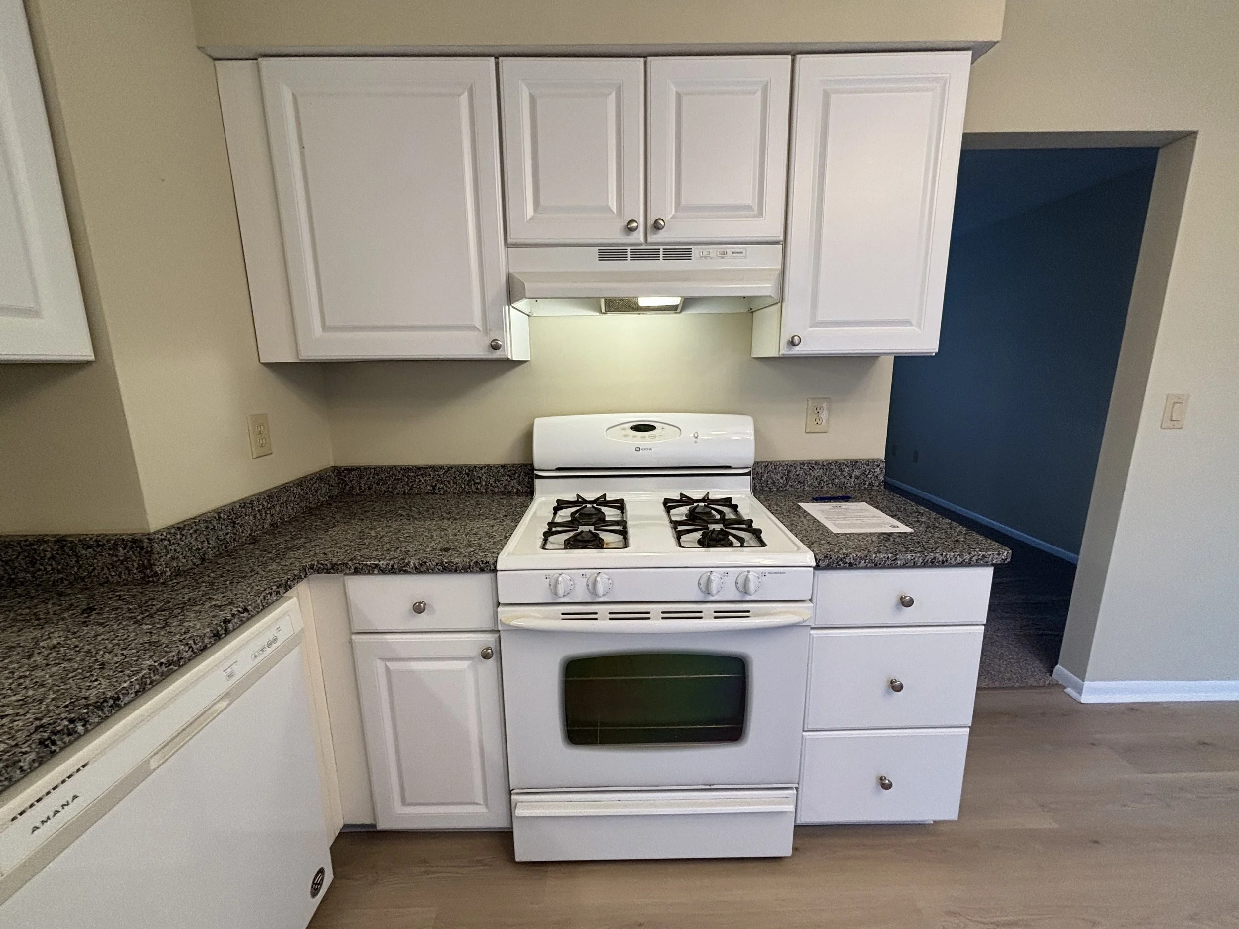 White kitchen stove with four burners and oven, surrounded by granite countertops and white cabinets, with a blue wall and beige wall in the background.