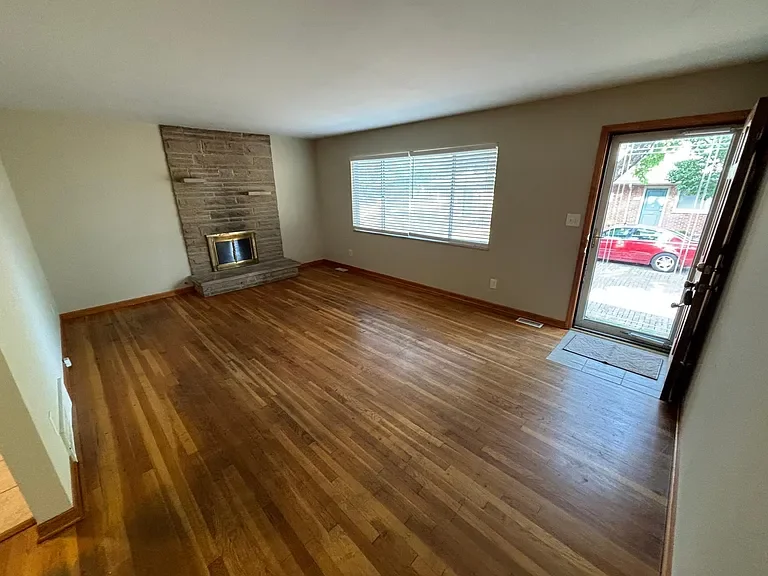 Empty living room with hardwood floors, a brick fireplace, a large window with blinds, and a glass door leading outside.