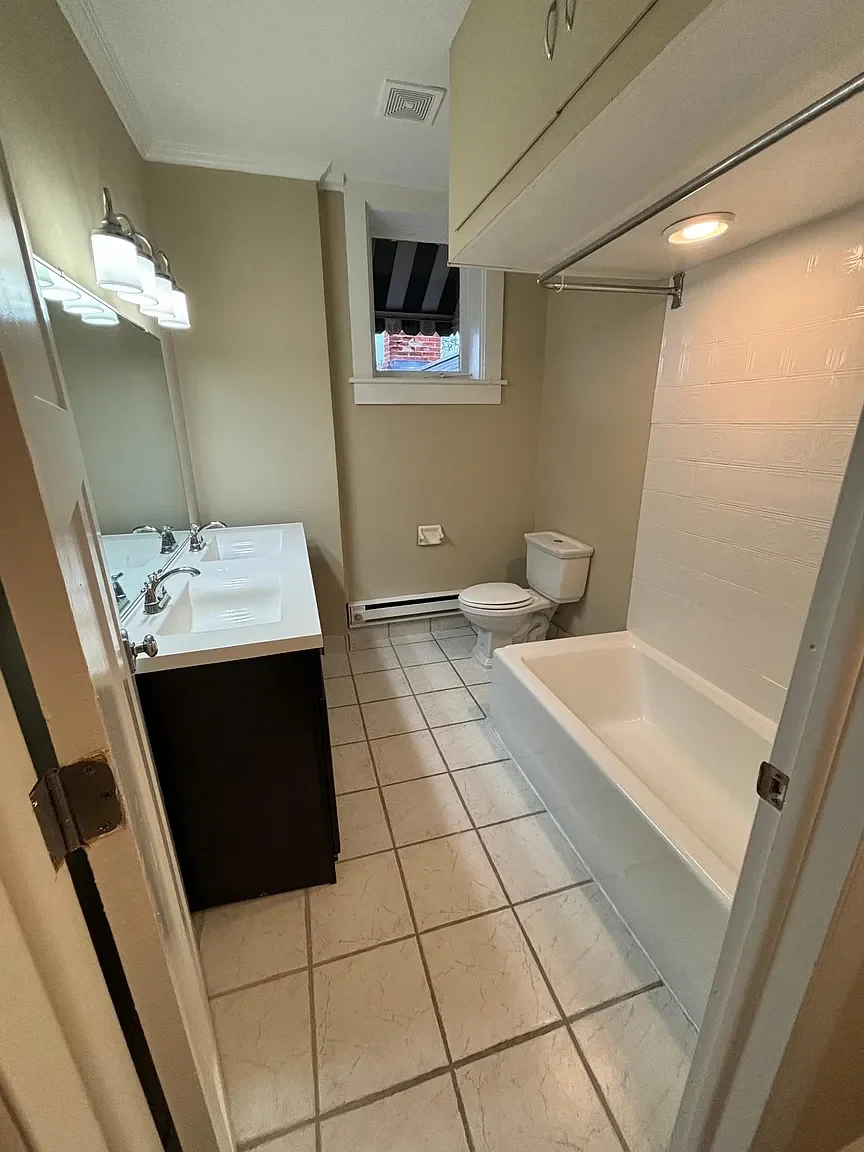 Bathroom with a double sink vanity, a toilet, a bathtub, a window, and tiled floor.