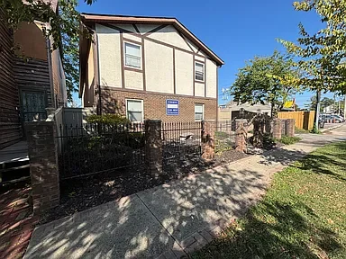 A two-story residential house with beige siding and brick foundation, situated on a sidewalk with a grassy yard and trees.